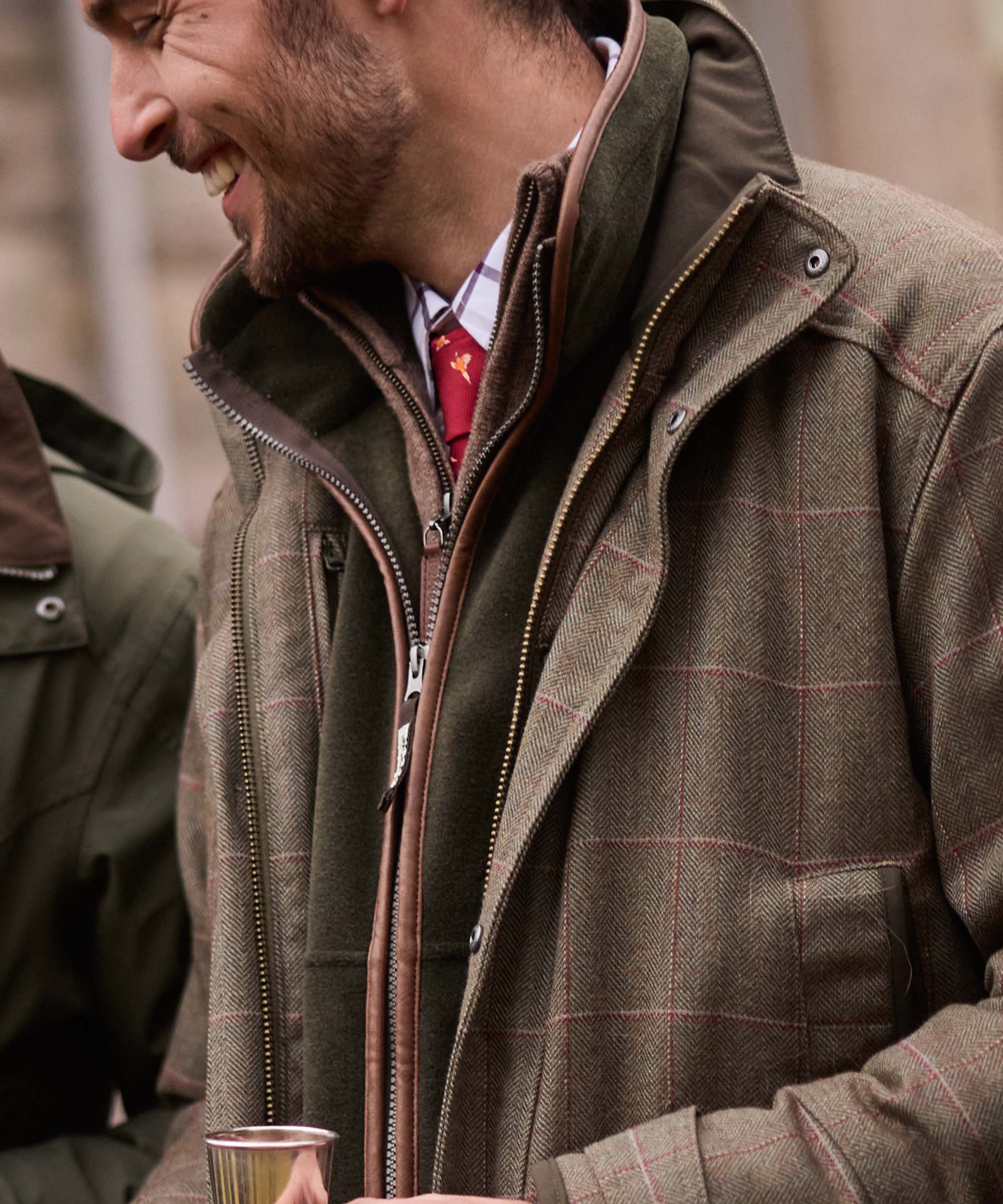 Close-up of an olive tweed shooting coat's leather-trimmed zip and stand collar, layered over a green fleece and check shirt with red tie