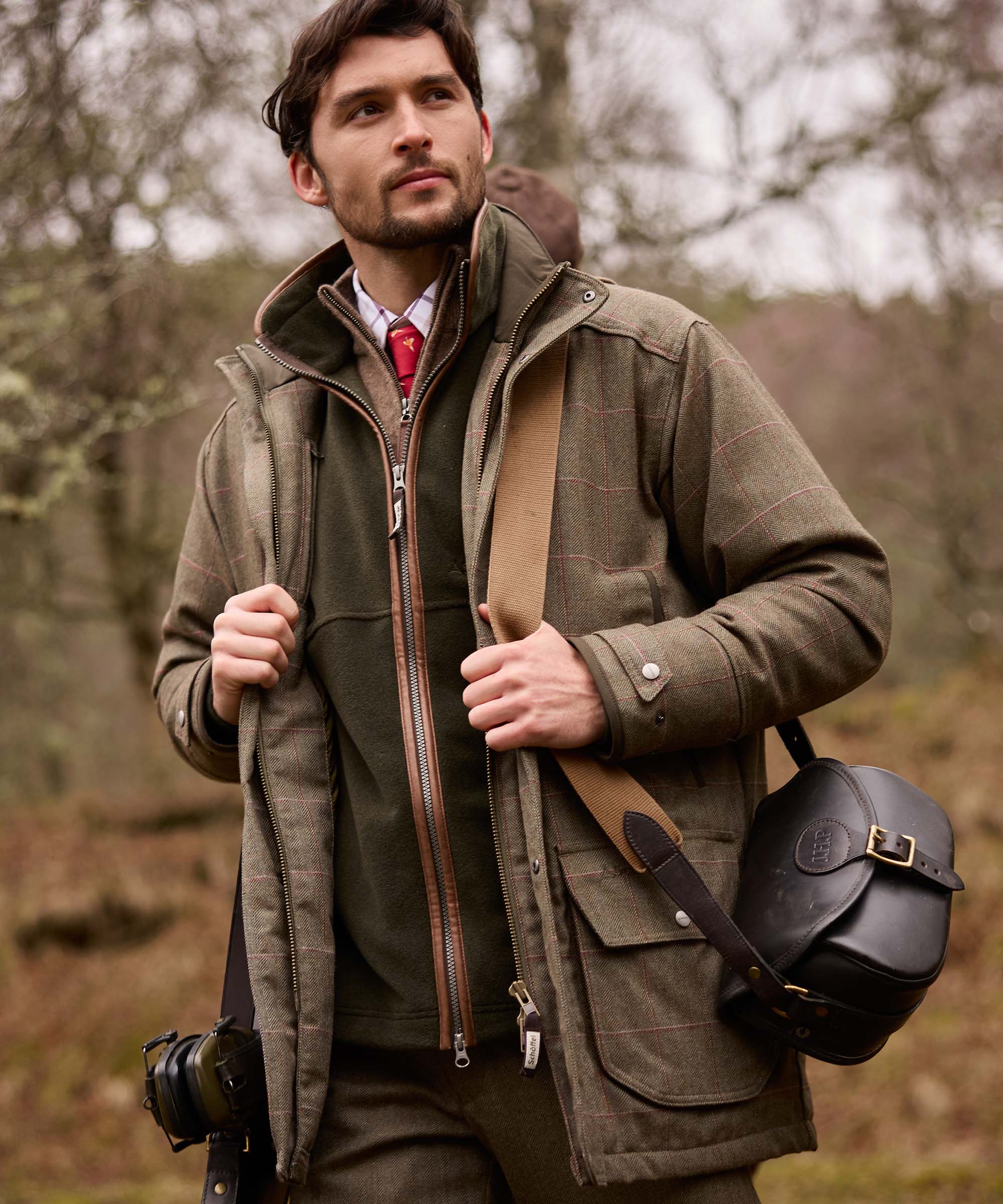 Man adjusting a leather cartridge bag strap, wearing an olive tweed shooting coat with corduroy-lined hood and multiple bellows pockets