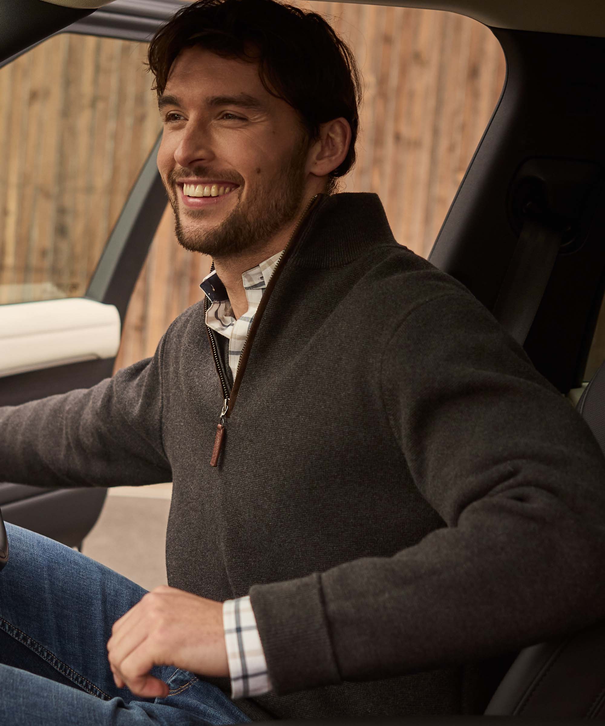 Man smiling behind the wheel wearing a charcoal quarter-zip jumper with leather zip pull, layered over a check shirt