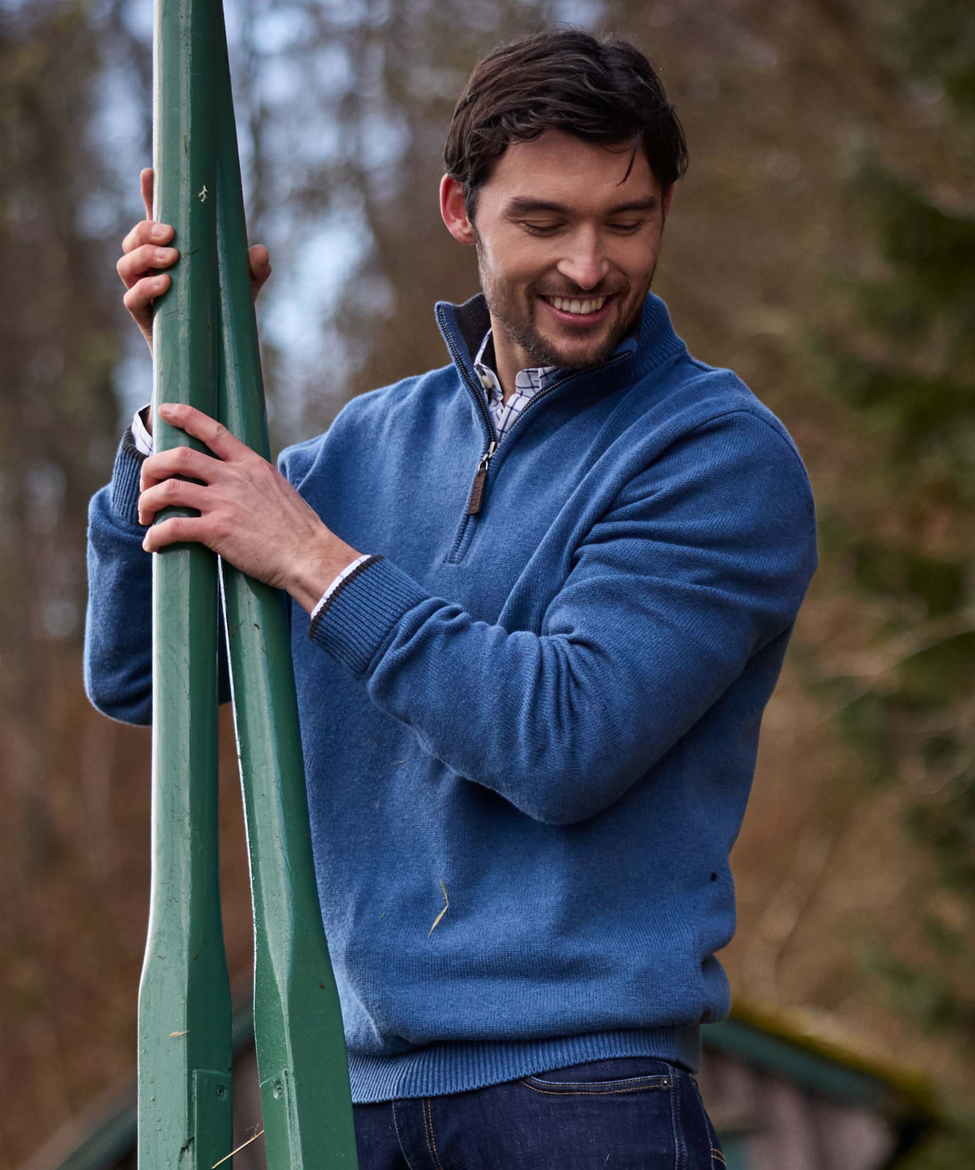 Man holding an oar smiling, wearing a denim-blue quarter-zip jumper with ribbed cuffs