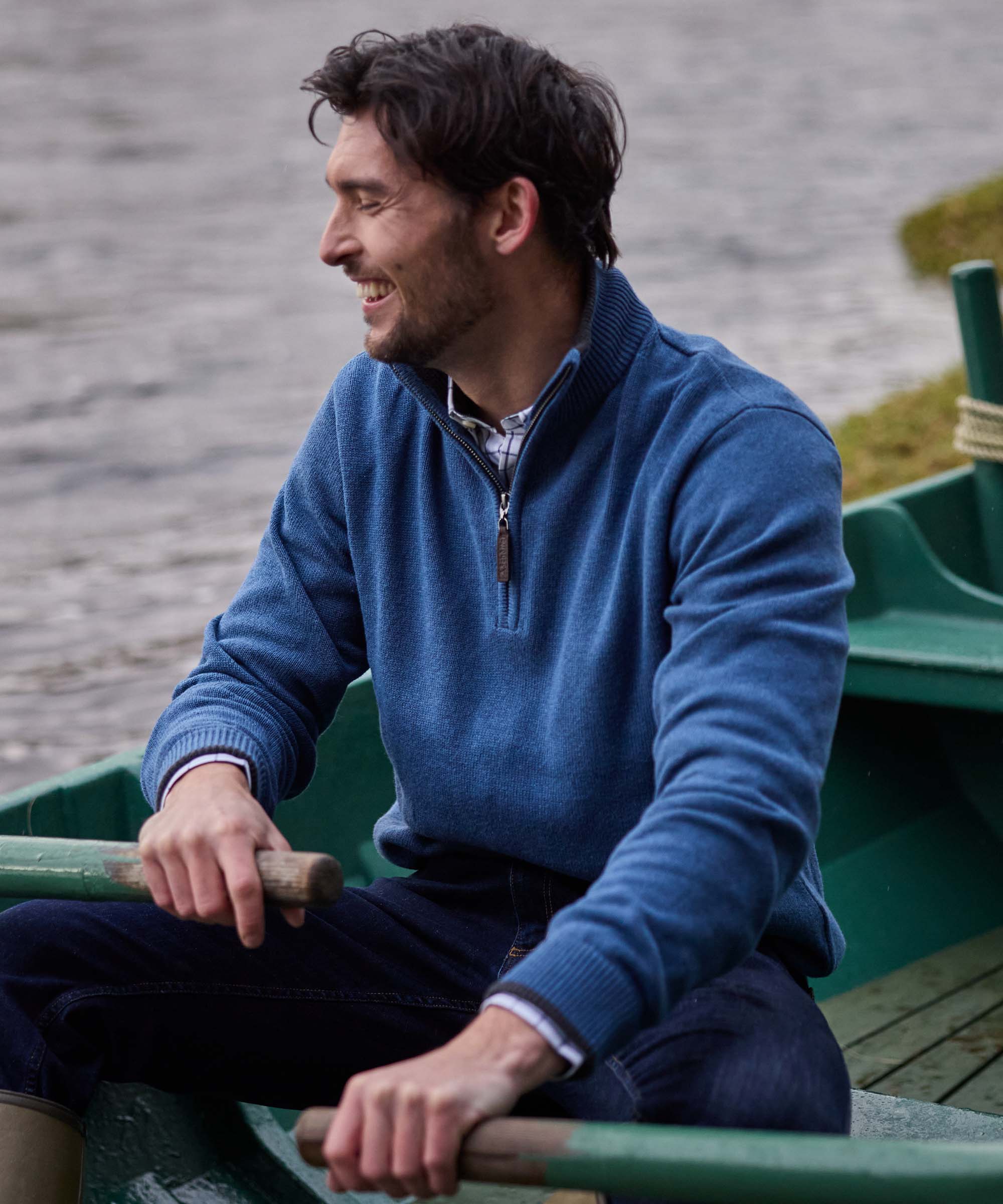 Man laughing while rowing a boat, wearing a denim-blue quarter-zip jumper over a check shirt