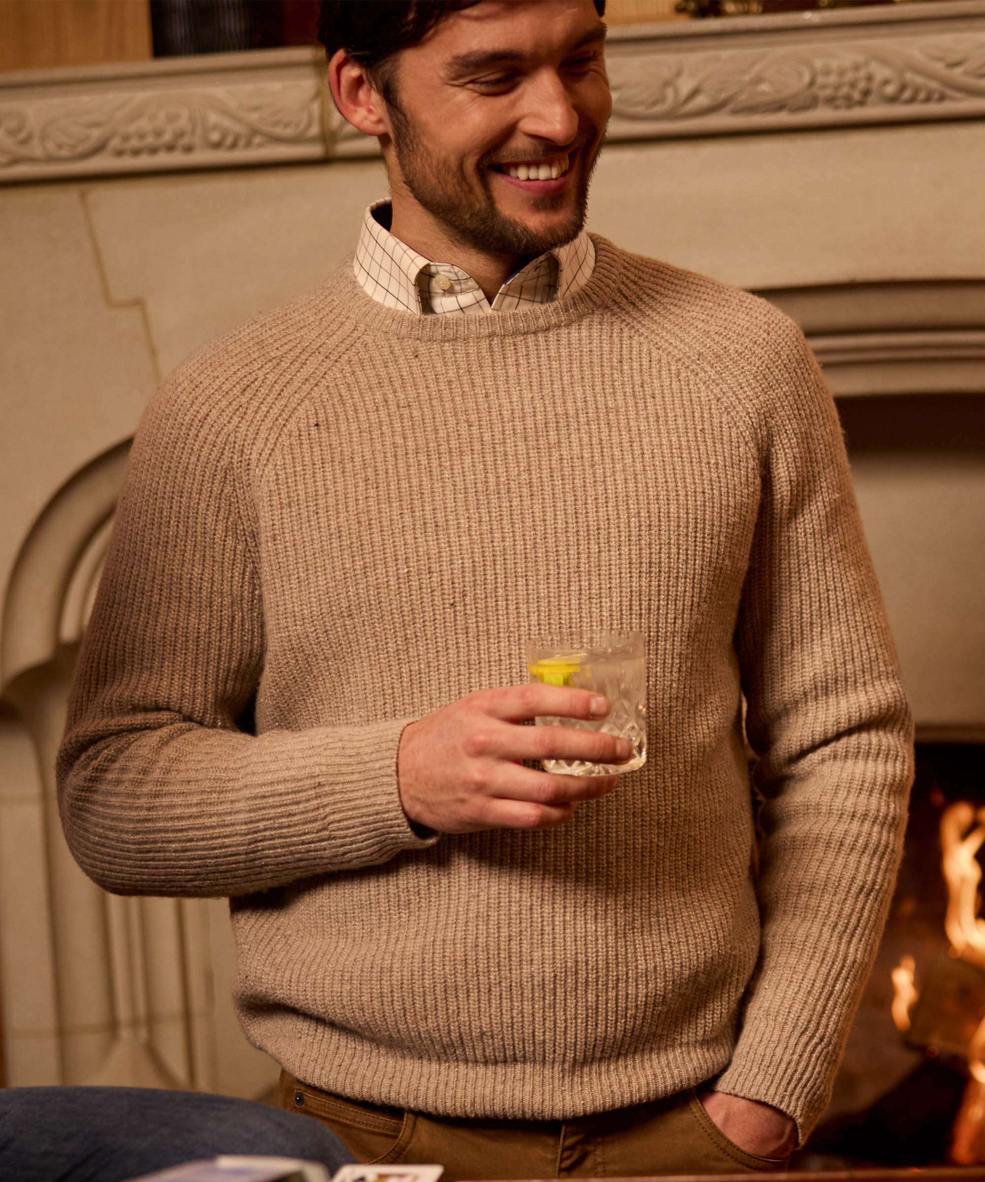 Man smiling by a fireplace holding a drink, wearing an oatmeal ribbed crew neck jumper over a check shirt