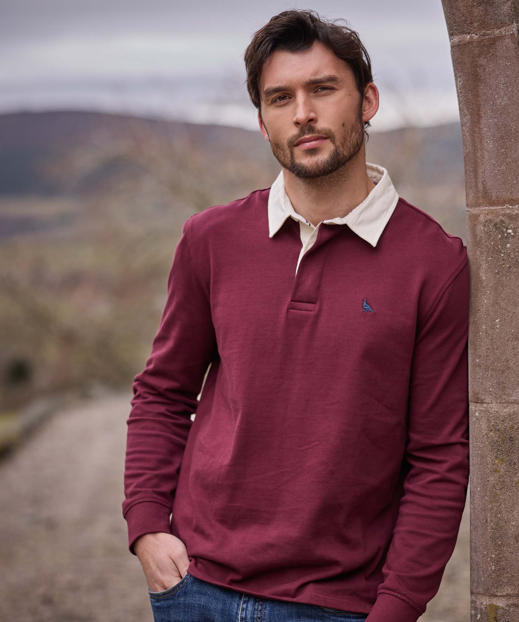 Man leaning against a stone pillar wearing a burgundy rugby shirt with a cream contrast collar and embroidered game bird motif