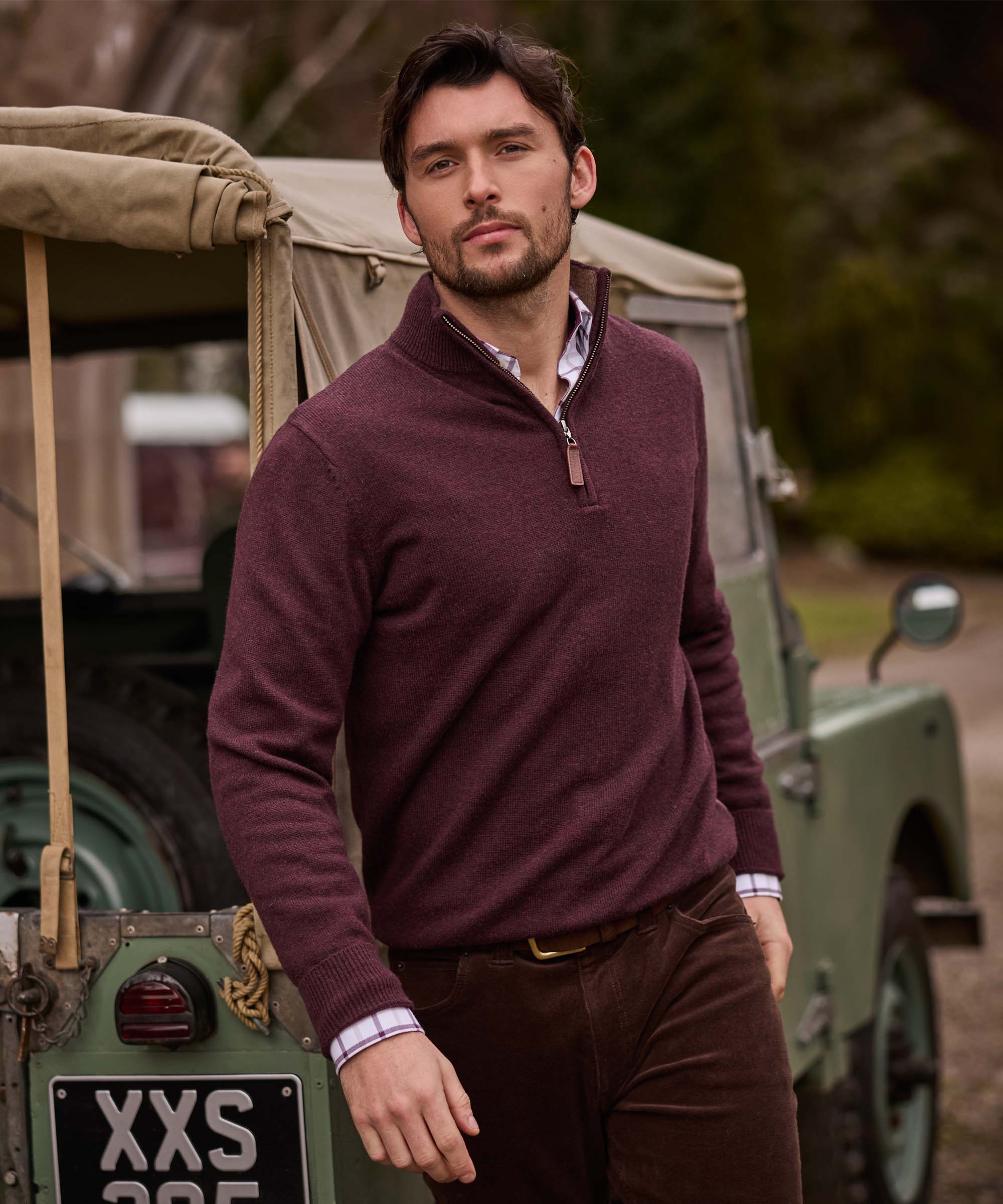 Man standing beside a vintage Land Rover wearing a burgundy quarter-zip jumper with brown corduroy trousers