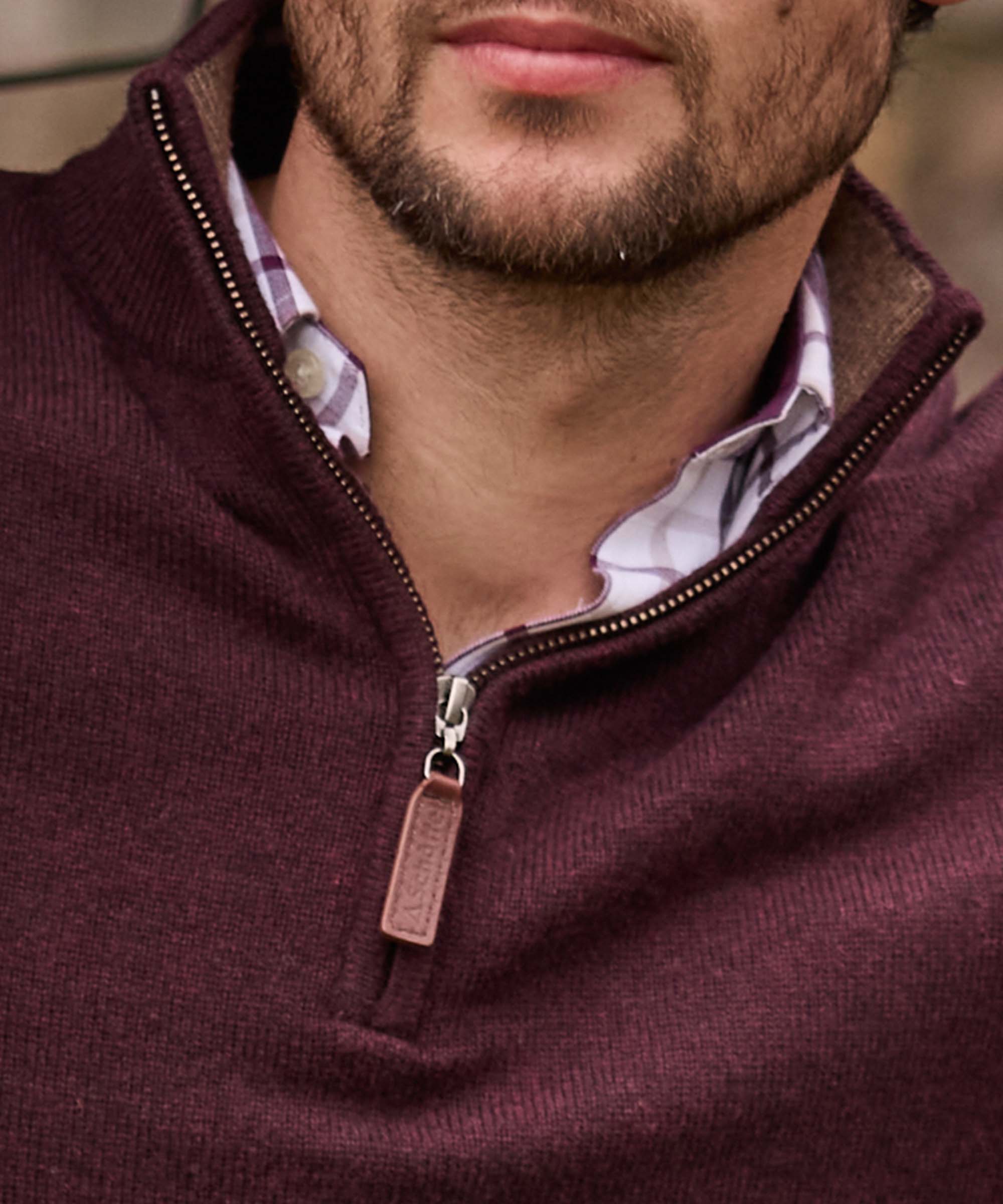 Close-up of a burgundy quarter-zip jumper's leather-trimmed zip, layered over a checked shirt