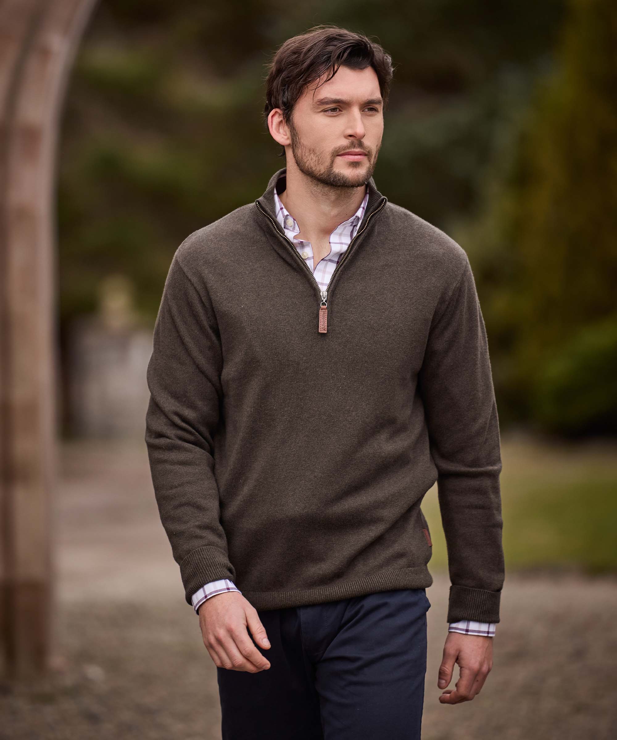 Man walking outdoors wearing an olive quarter-zip jumper over a check shirt, paired with navy trousers