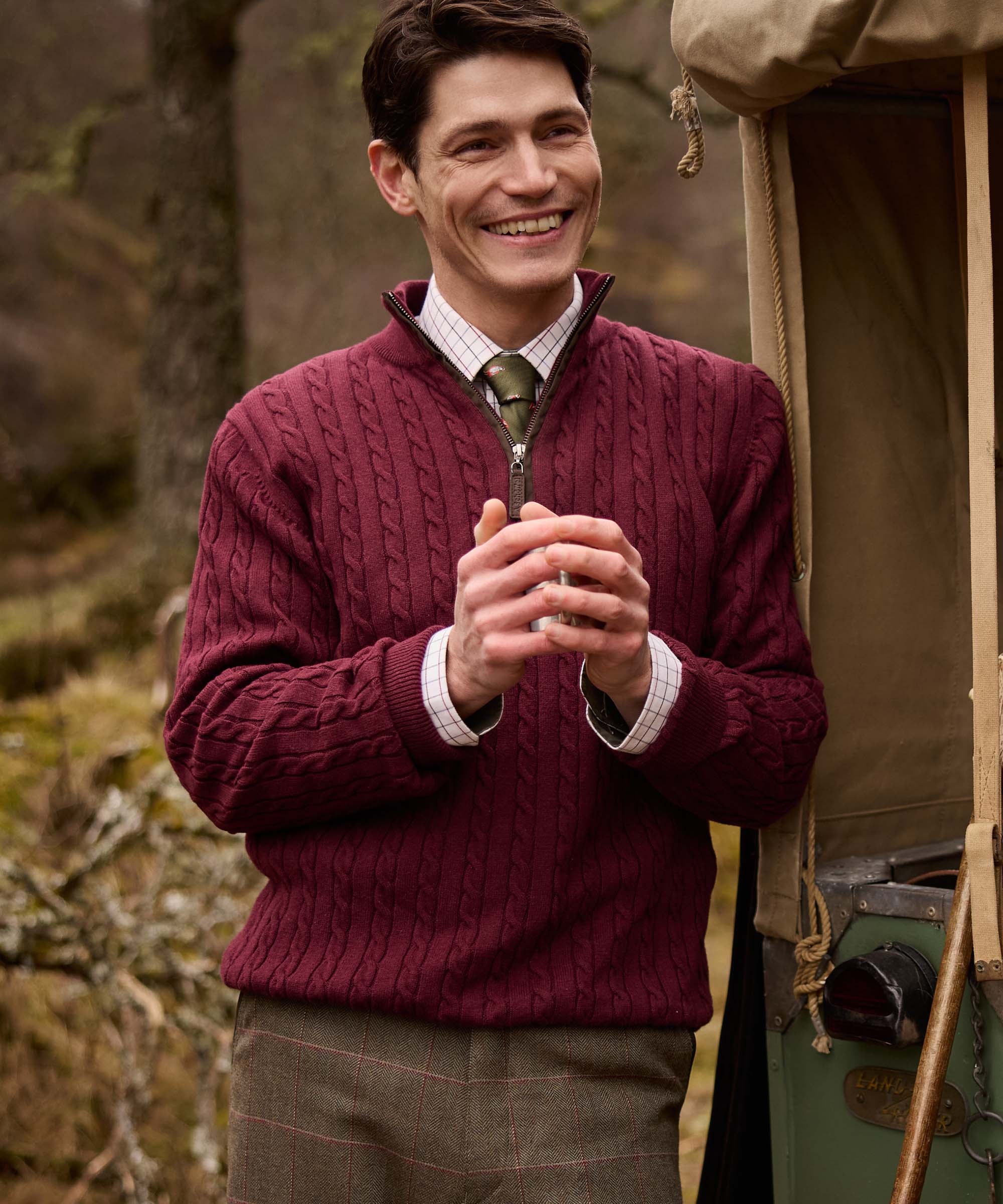 Man smiling and holding a flask cup, wearing a burgundy cable knit quarter-zip jumper with a check shirt and olive tie, styled with tweed trousers