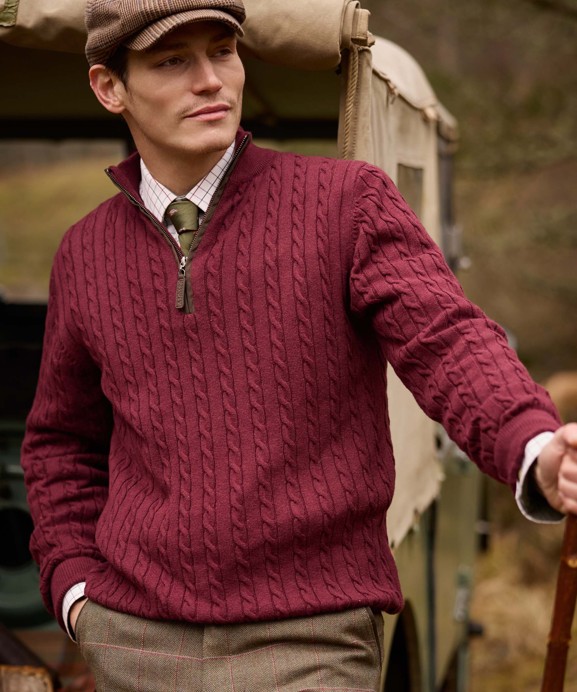 Man in a herringbone flat cap wearing a burgundy cable knit quarter-zip jumper over a check shirt and tie