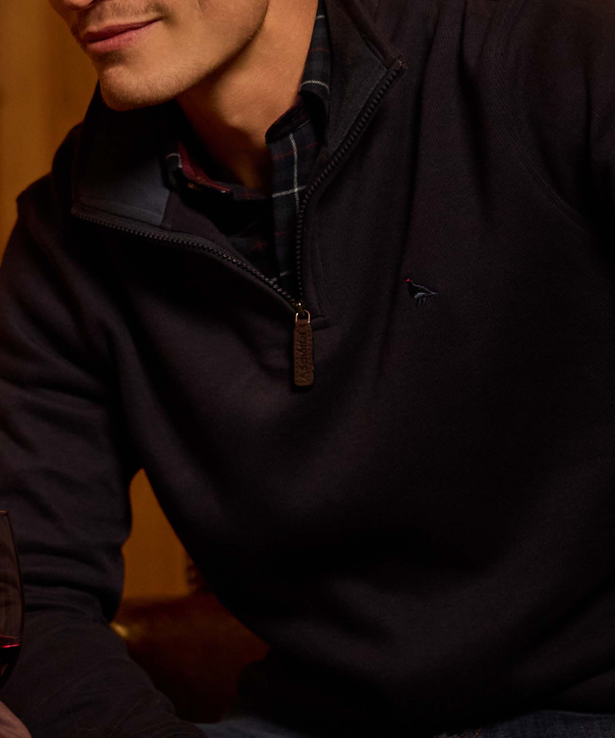 Close-up of a man in a navy quarter-zip jumper with embroidered game bird motif, worn over a tartan-collared shirt