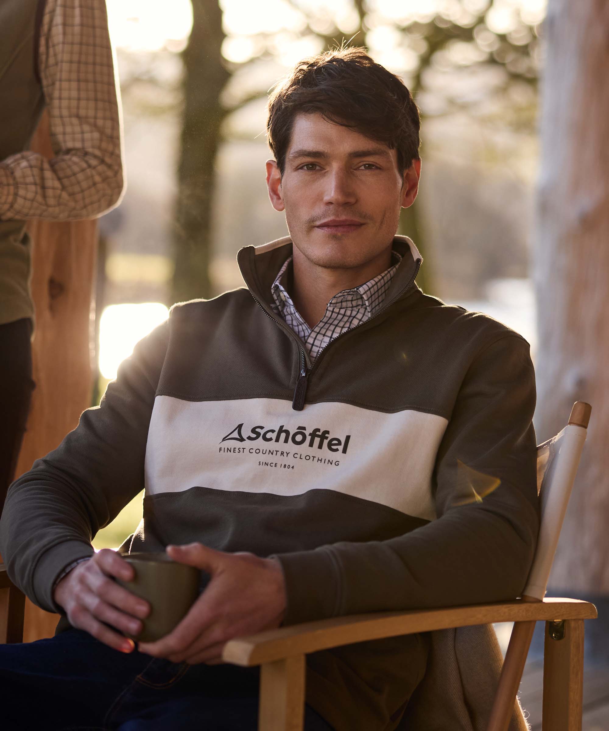 Man seated outdoors holding a mug, wearing a khaki and cream colour-block quarter-zip jumper with Schöffel branding