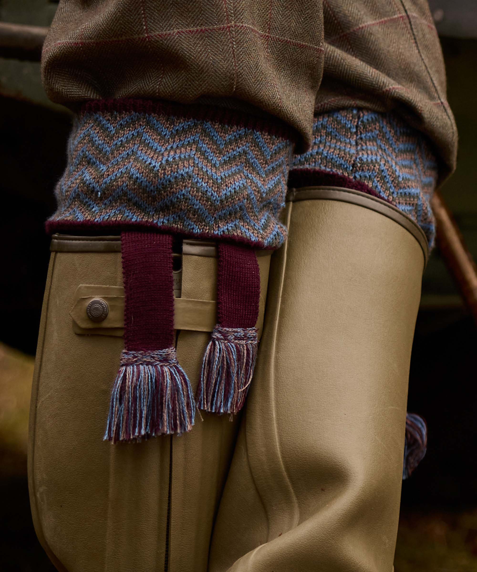 Close-up of matching Fair Isle sock garters with burgundy tasselled ties, worn over tweed breeks and leather wellington boots
