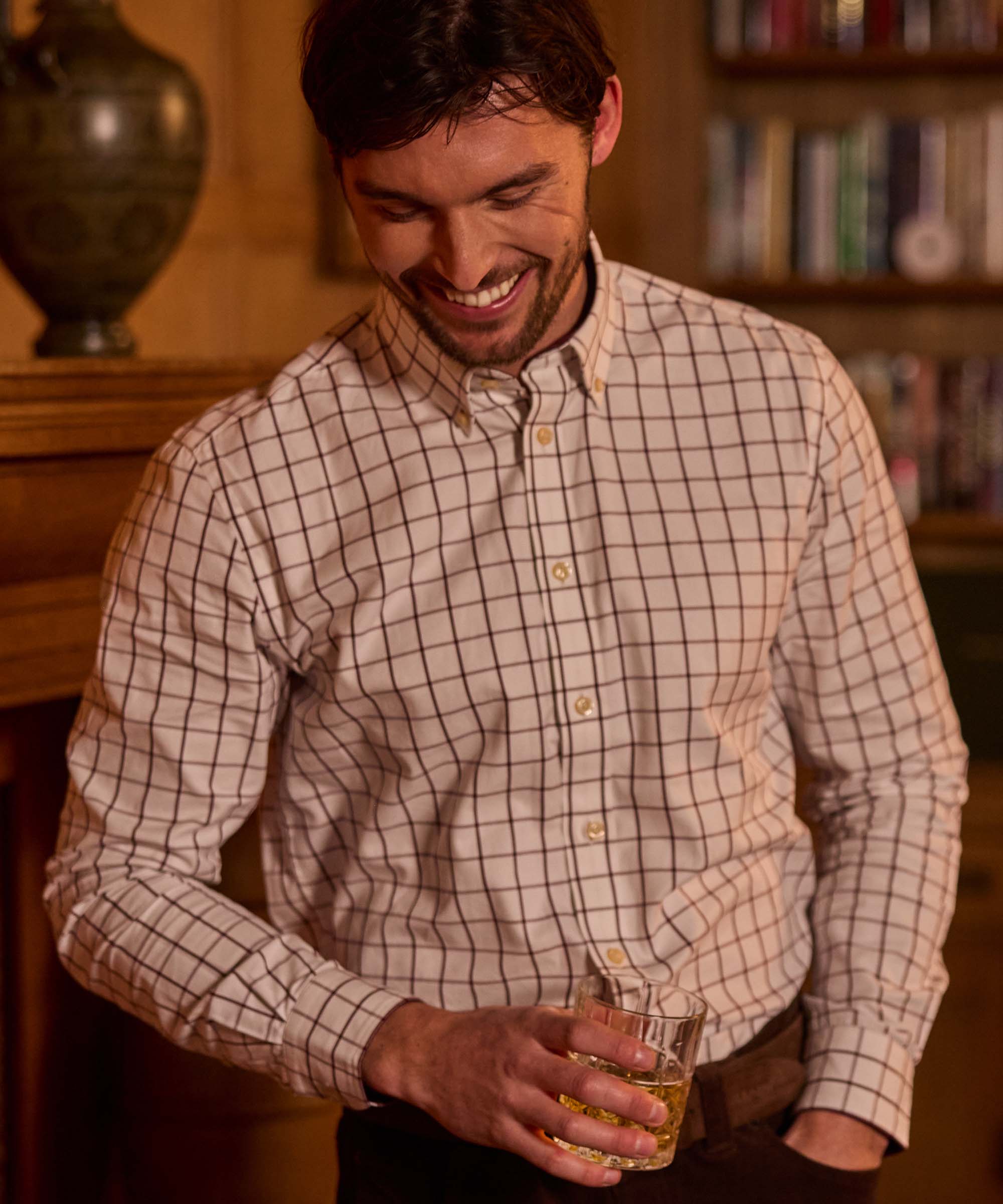 Man smiling and holding a whisky glass, wearing a white and burgundy windowpane check shirt