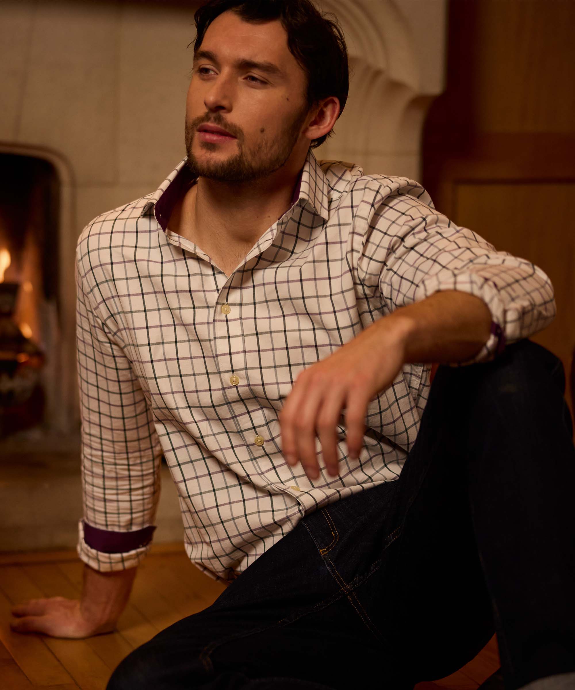 Man sitting by a fireplace wearing a cream check shirt with a contrasting burgundy trim inside the collar and cuffs