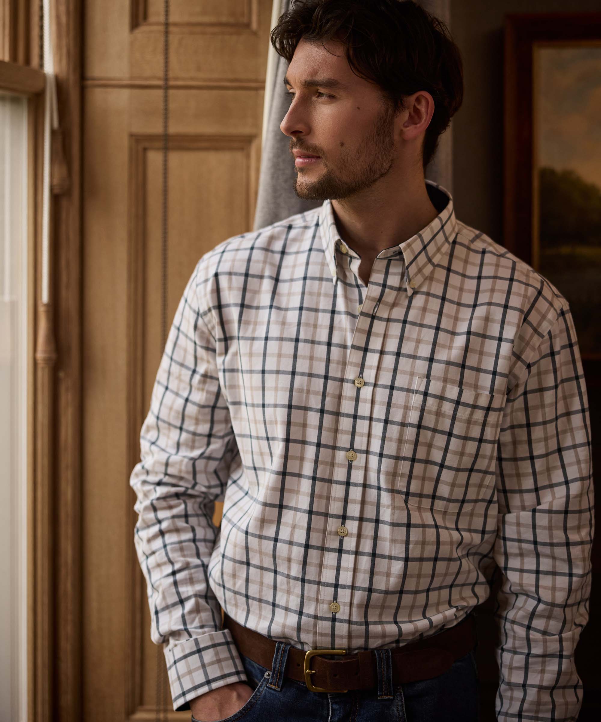 Man standing by a window wearing a beige and navy check shirt with button-down collar and chest pocket