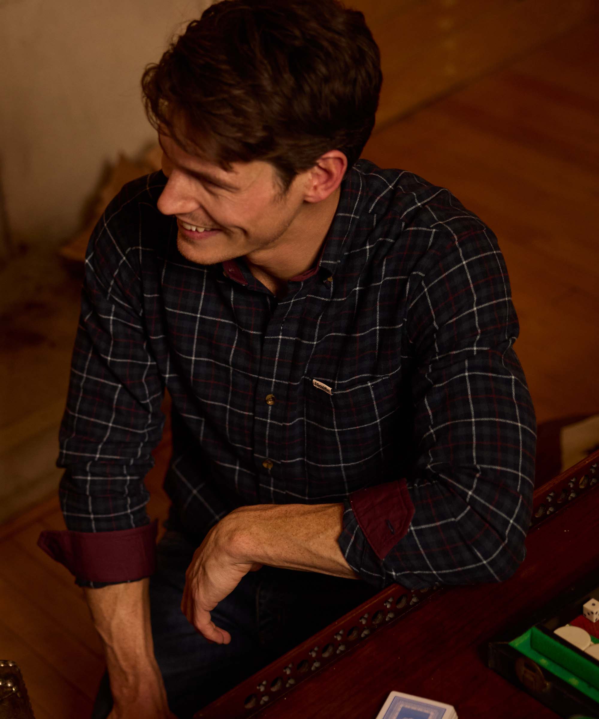 Man smiling over a backgammon board wearing a navy check flannel shirt with contrasting burgundy cuff and collar trim