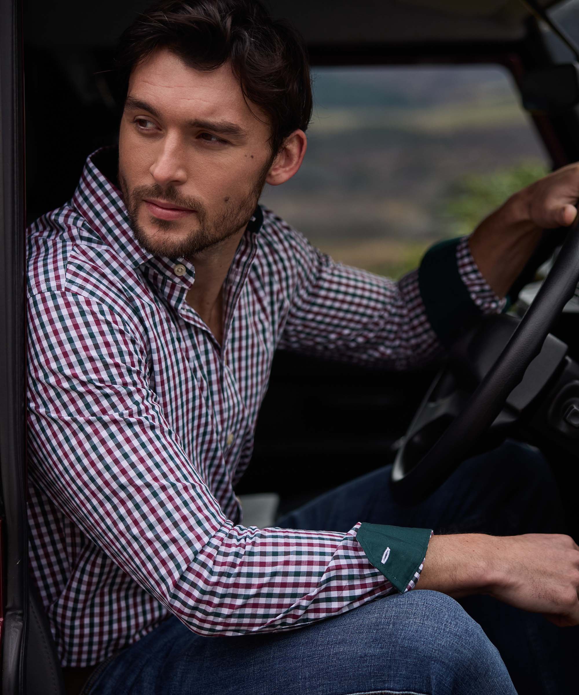 Man driving with one hand on the wheel, wearing a burgundy, green and white gingham check shirt