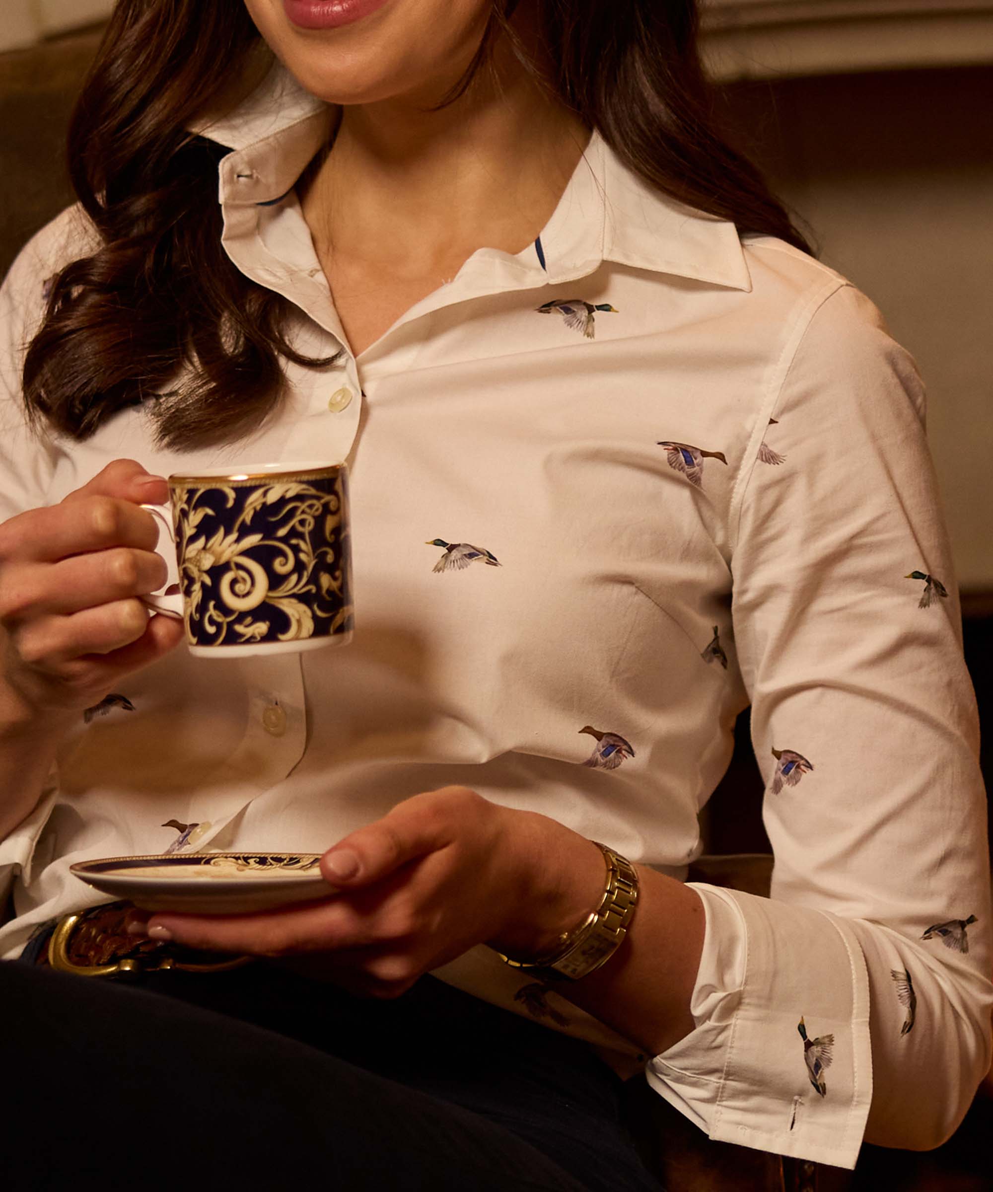 Close-up of a woman holding a teacup and saucer, wearing a white mallard duck-print shirt with a gold watch