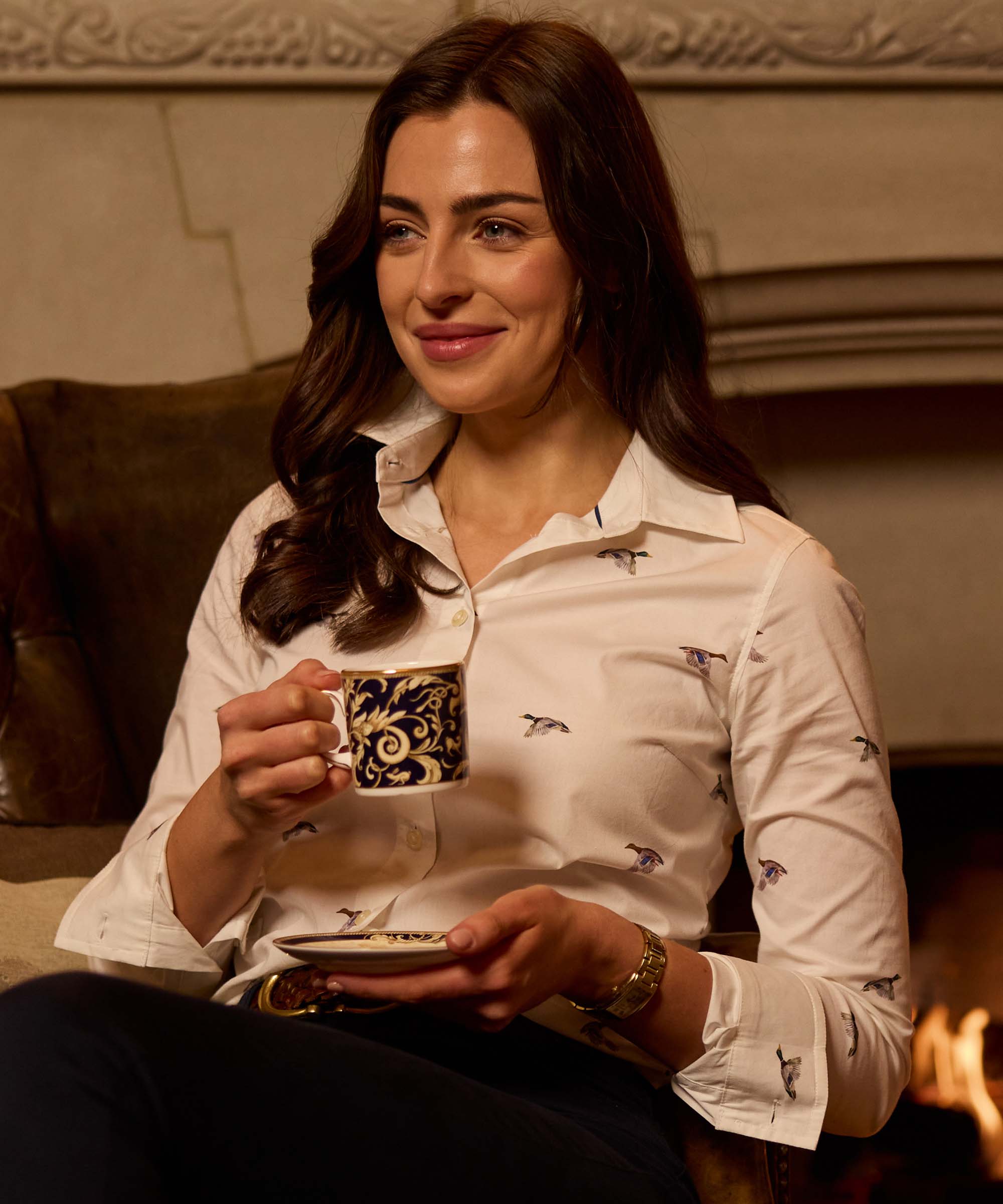 Woman holding a teacup by the fireside, wearing a white shirt with an all-over flying mallard duck print