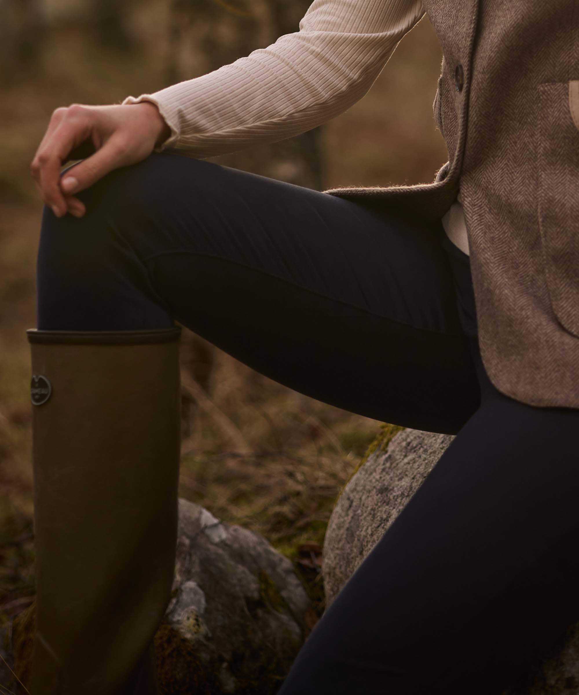 Close-up of a woman seated on a rock wearing navy technical riding trousers with a tweed gilet and wellington boots