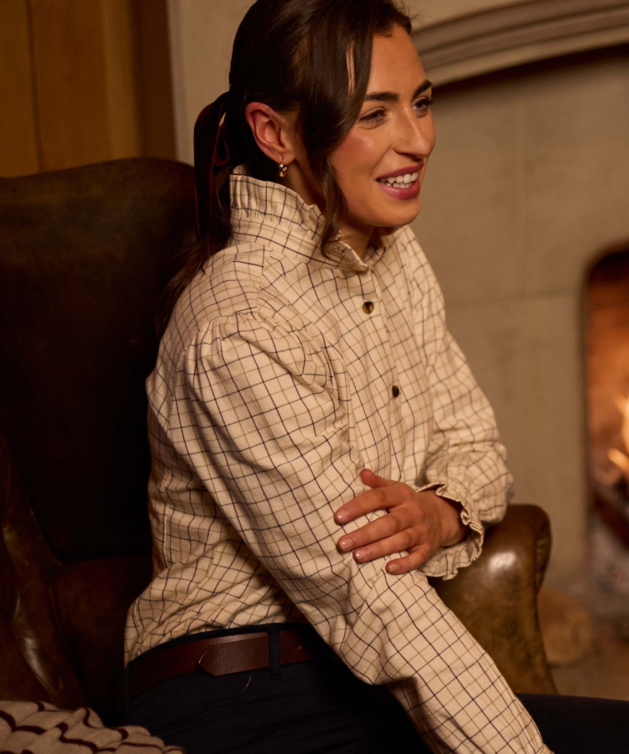 Woman wearing Schöffel Country's ivory check ruffle-neck shirt with tortoiseshell buttons, smiling by a fireside setting