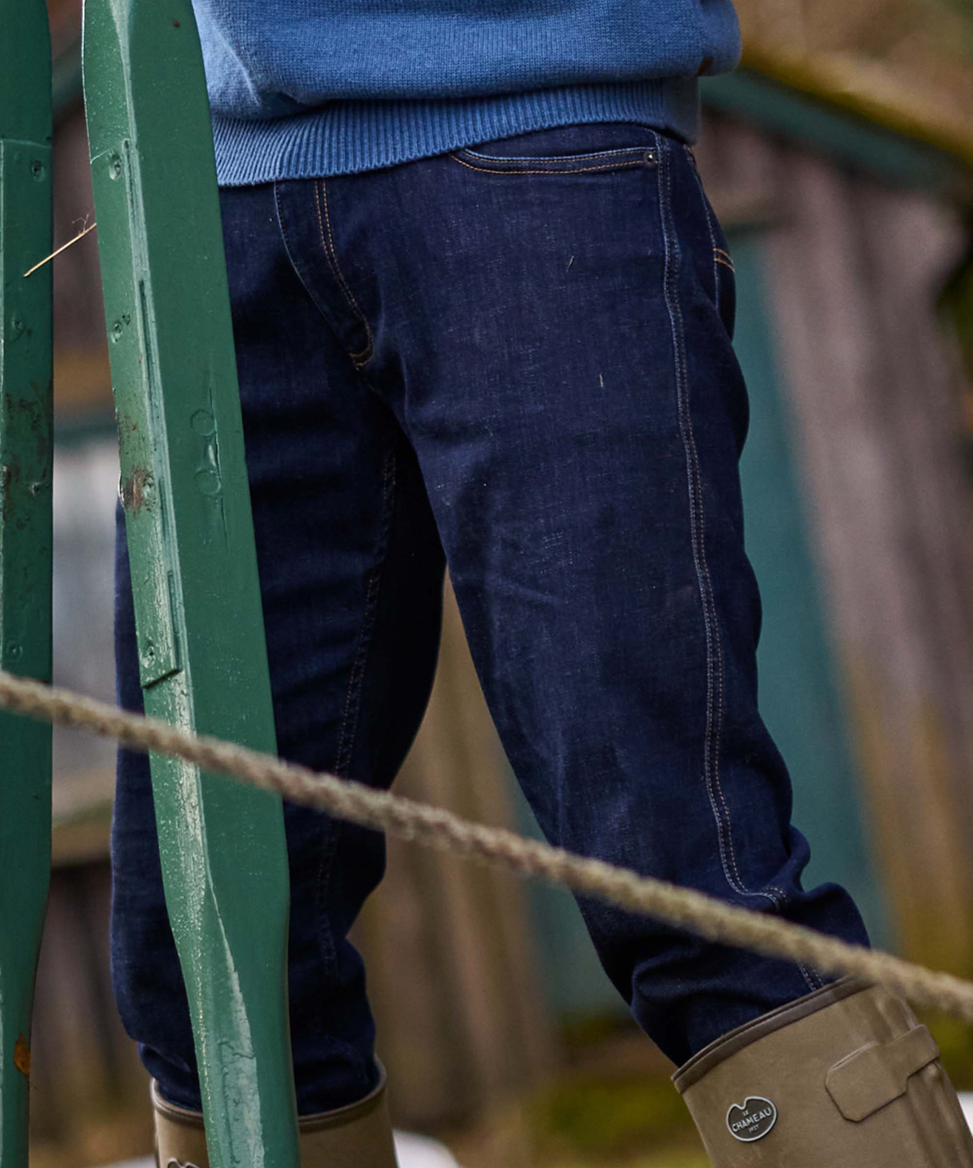 Close-up of a person wearing dark blue denim jeans, a blue top, and tan rubber boots, standing near a green metal structure with a rope in the foreground.