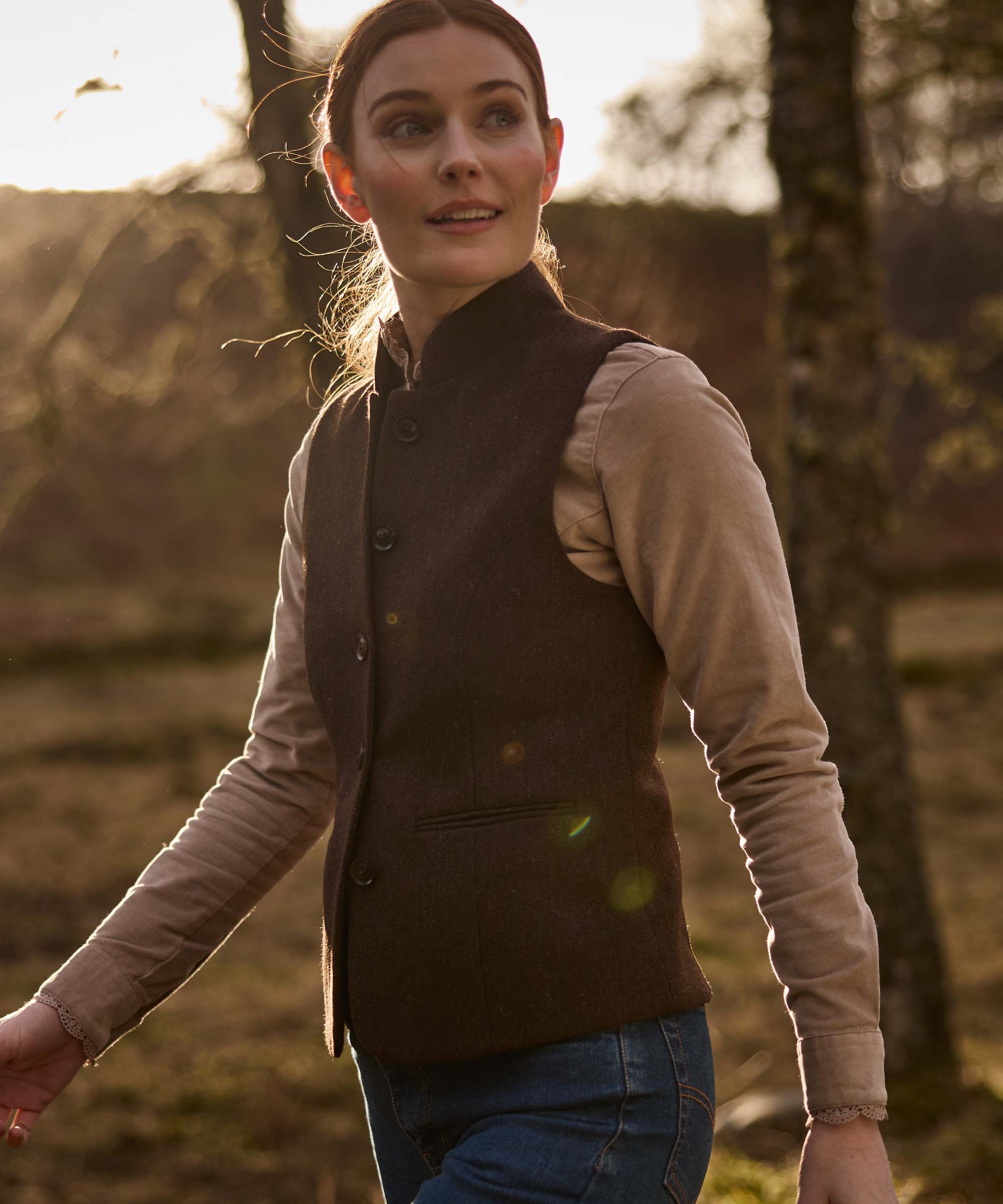 Woman wearing Schöffel Country's brown herringbone tweed gilet with stand collar, walking in woodland at dusk