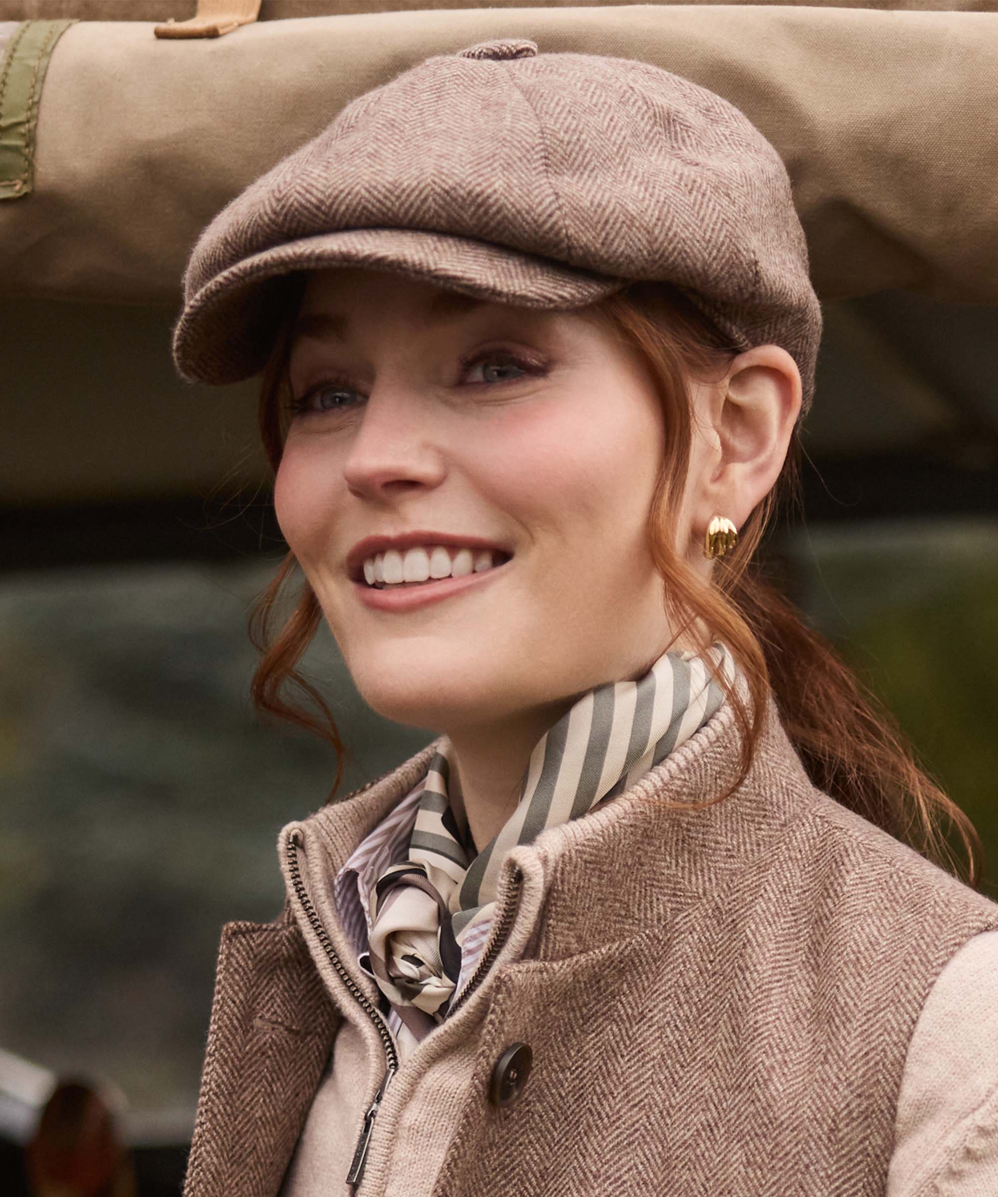 Woman wearing Schöffel Country's herringbone tweed flat cap with striped silk scarf, smiling beside a Land Rover