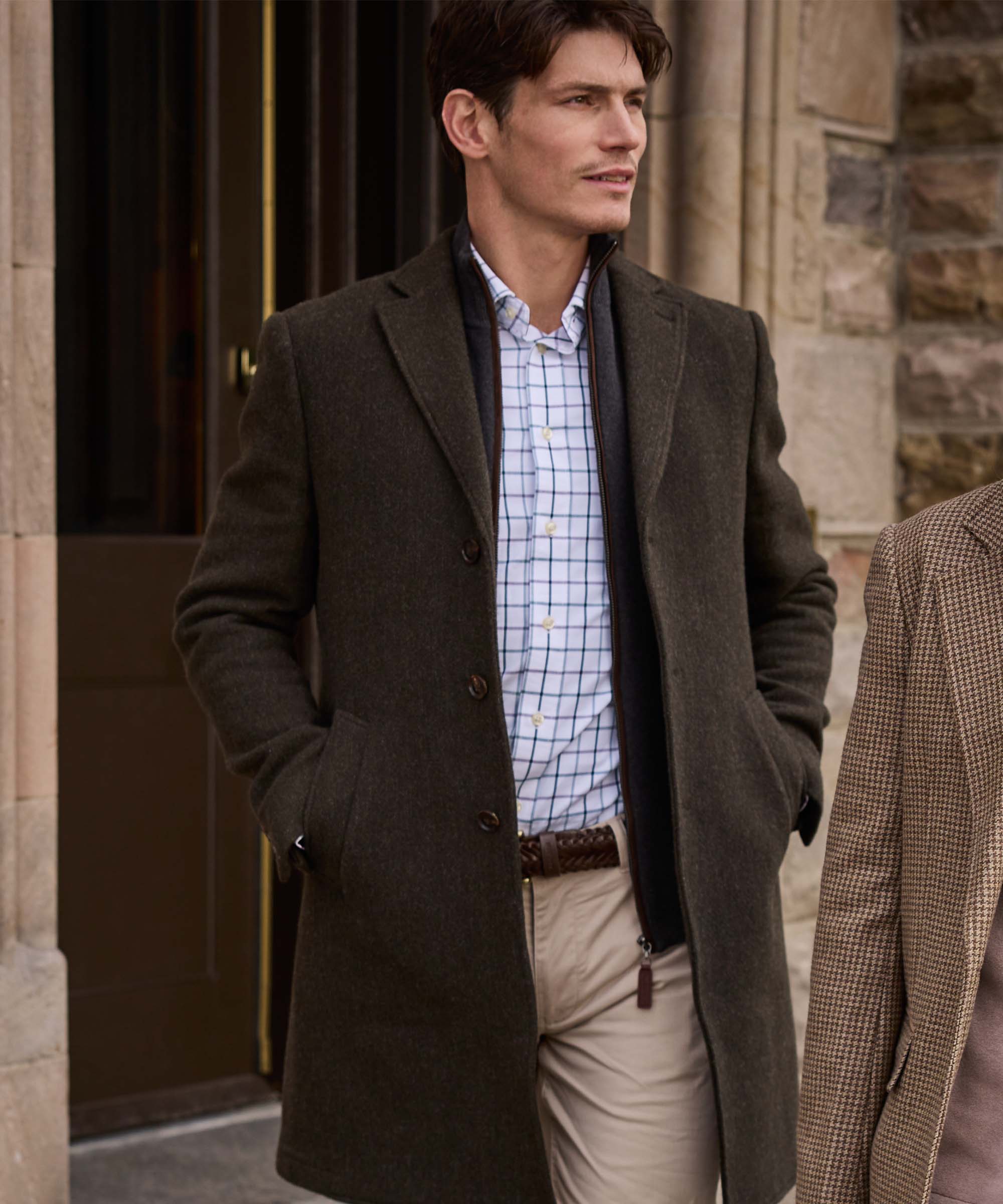 Man wearing Schöffel Country's khaki wool overcoat with check shirt and woven leather belt, stood outside a stone building