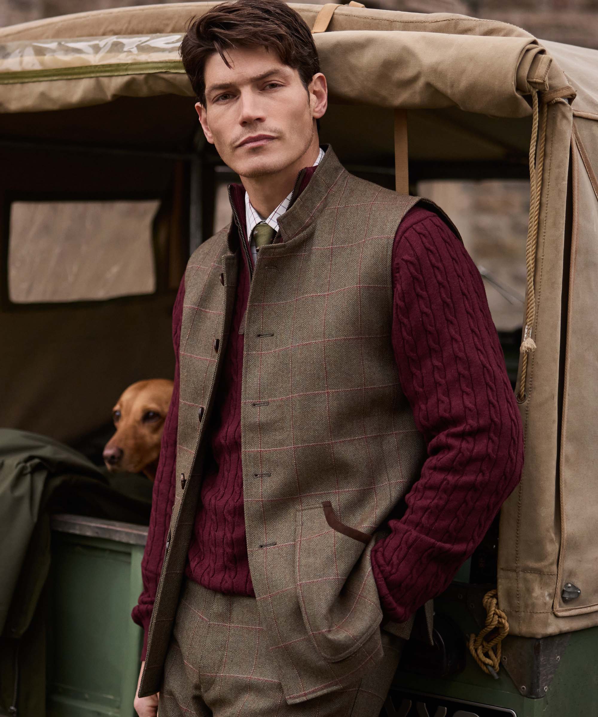 Man wearing Schöffel Country's tweed gilet with a burgundy cable knit jumper and check shirt, stood by a Land Rover with a Labrador