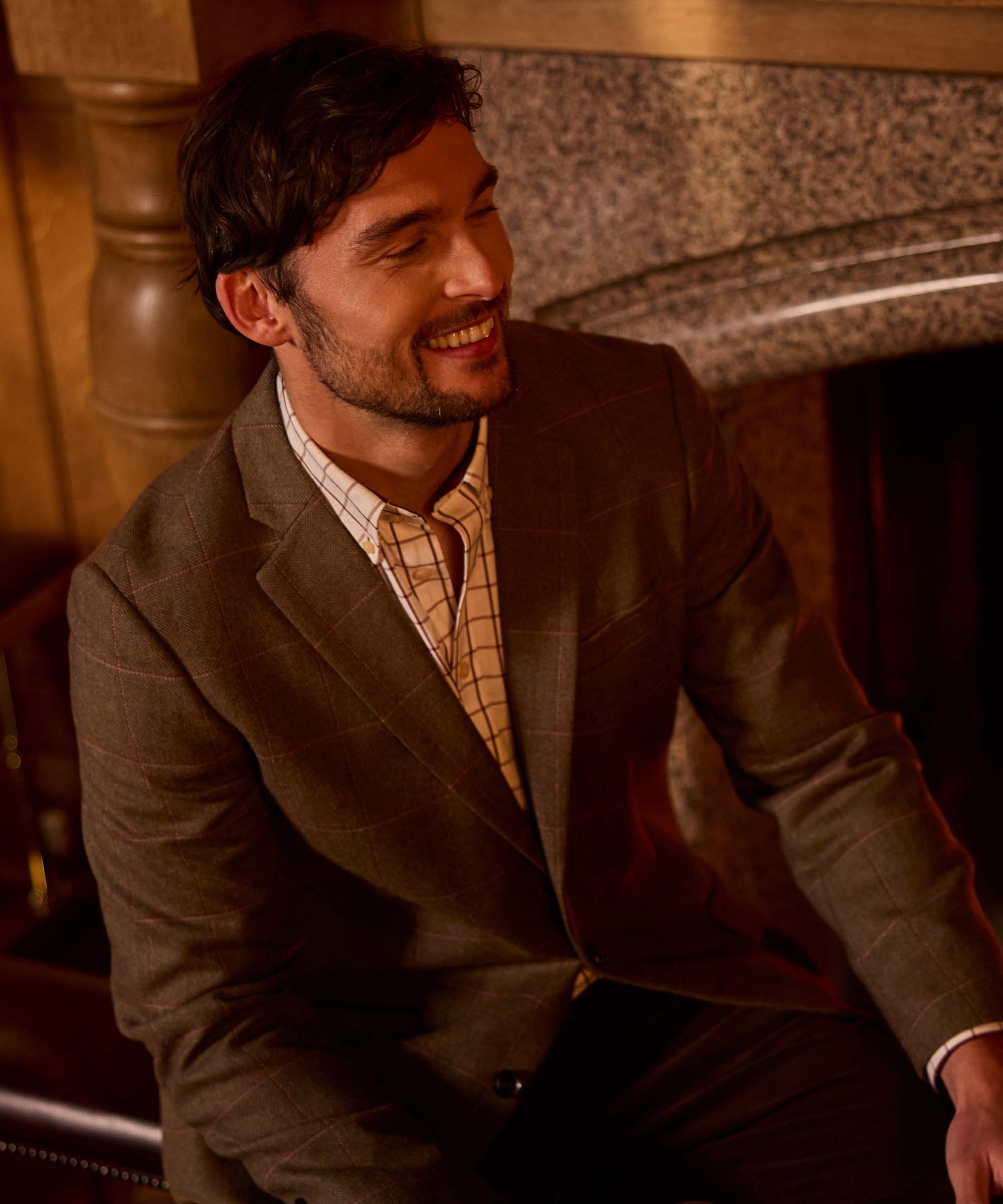 Man wearing Schöffel Country's brown herringbone tweed blazer with a check shirt, smiling by a fireplace