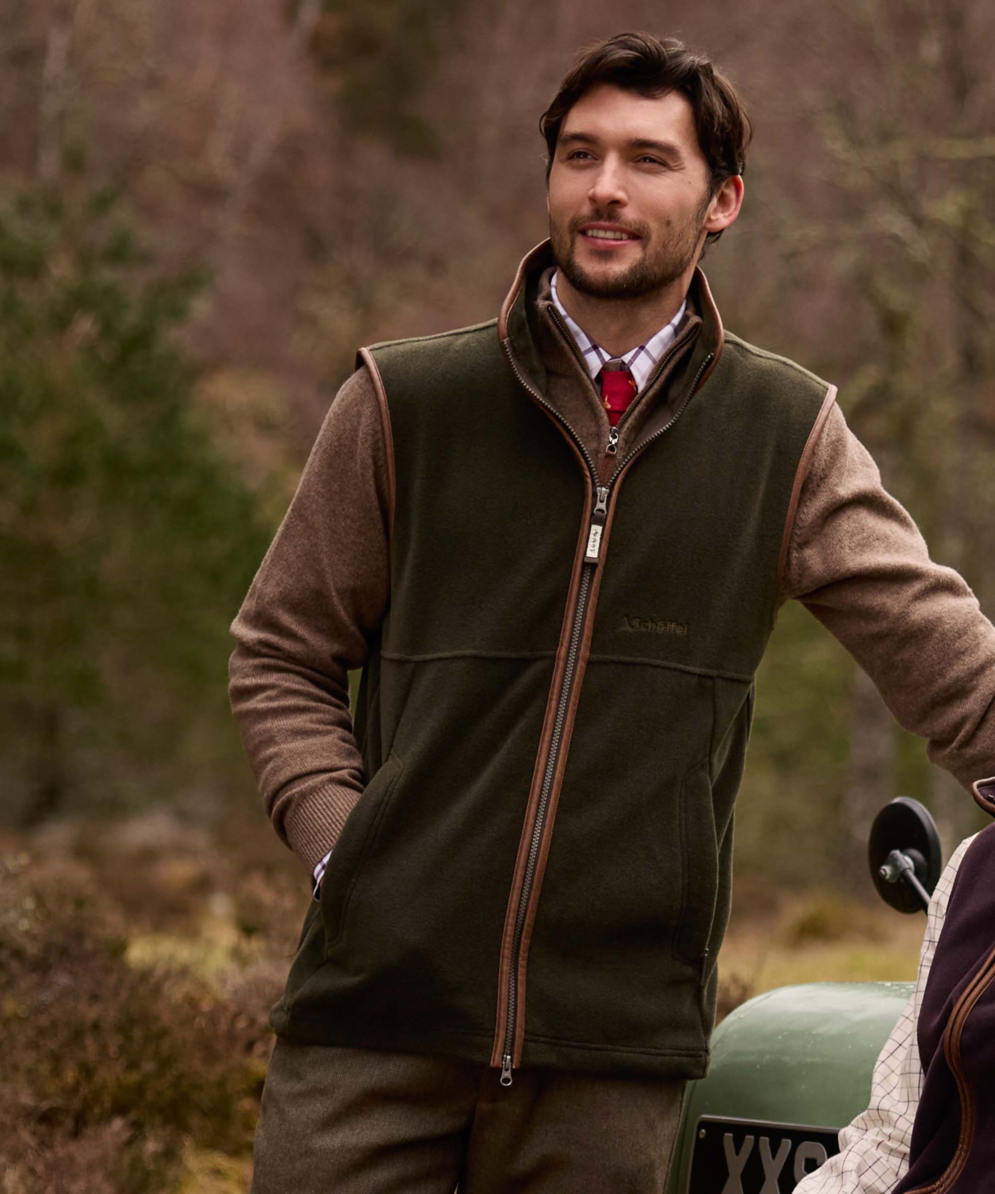 Man wearing Schöffel Country's khaki fleece gilet, smiling outdoors beside a Land Rover