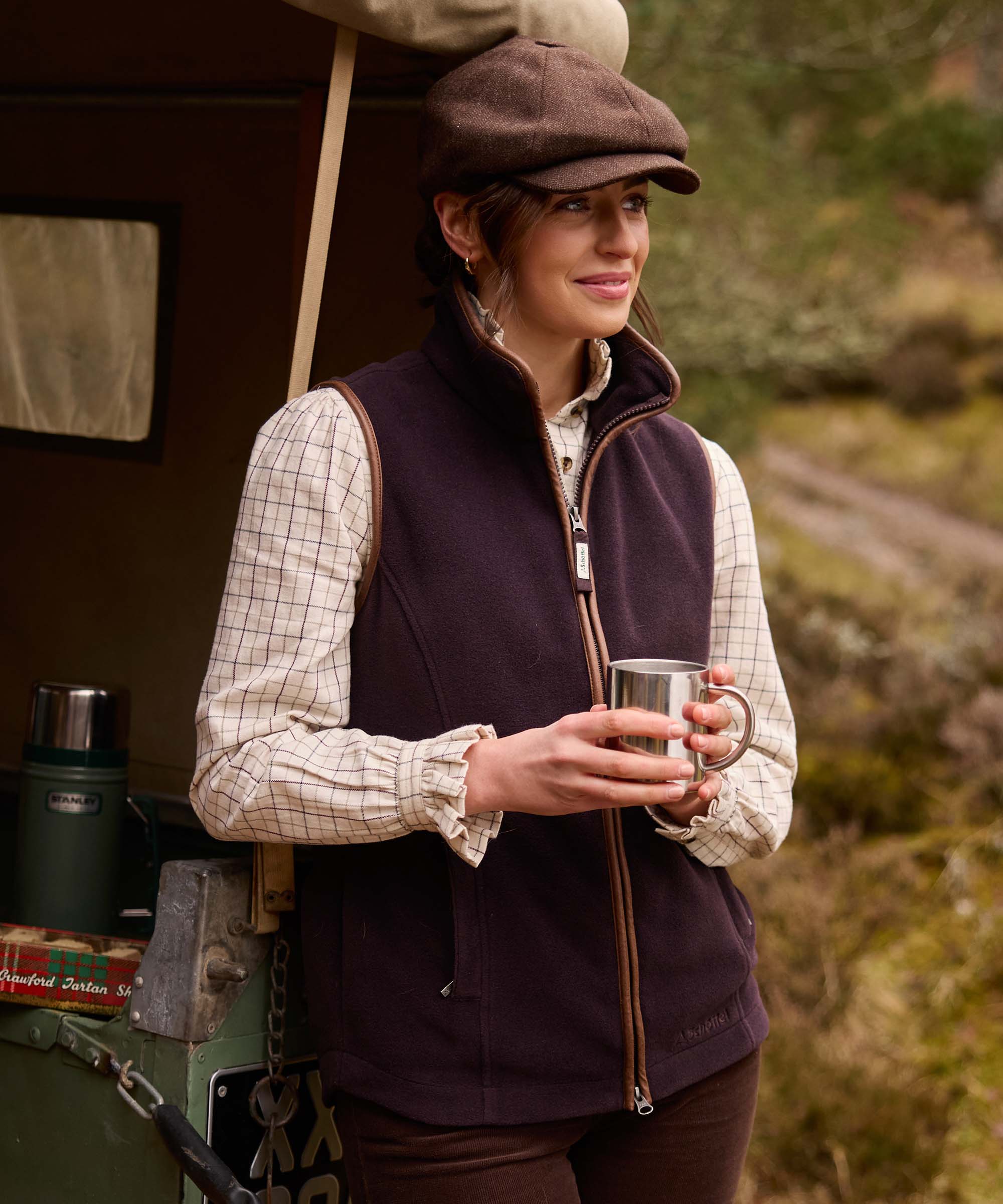 Woman wearing a plum Schöffel fleece gilet with brown corduroy trim over a checked shirt, holding a metal mug beside a Land Rover, wearing a tweed baker boy cap.