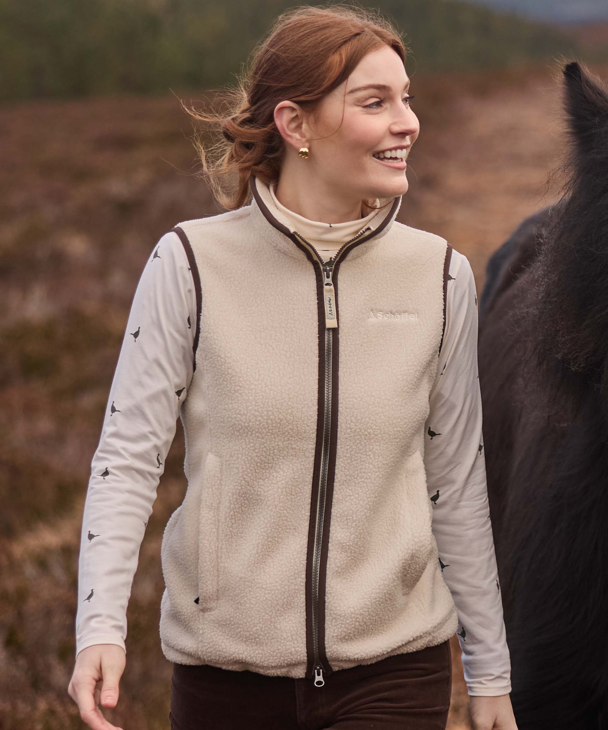Woman wearing a cream Schöffel sherpa fleece gilet over a pheasant-print top, smiling beside a horse in the Highlands.