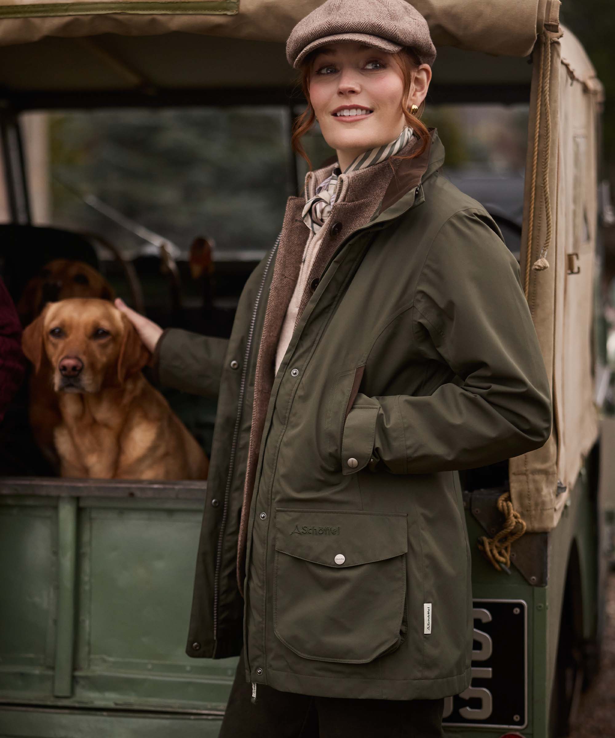 Woman wearing an olive green Schöffel waxed jacket and tweed cap, standing beside a Land Rover with a Labrador.
