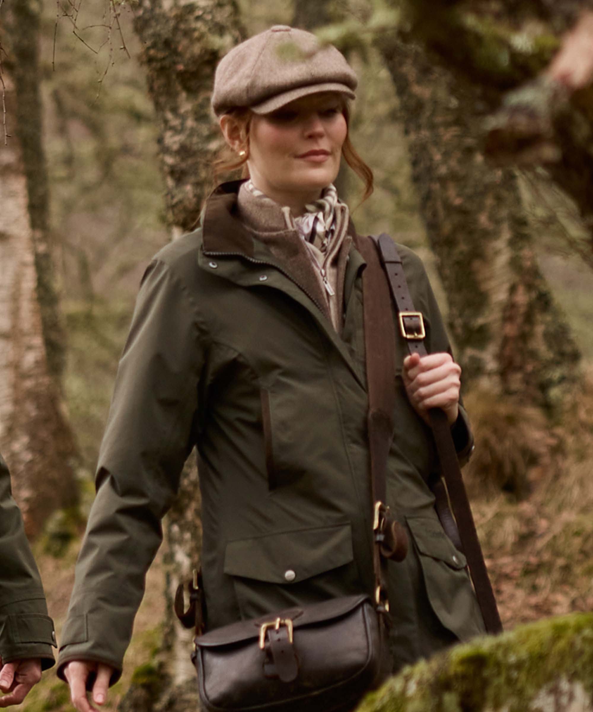 A woman wearing an olive green waterproof jacket, a brown crossbody bag, and a brown flat cap, standing in a mossy forest.