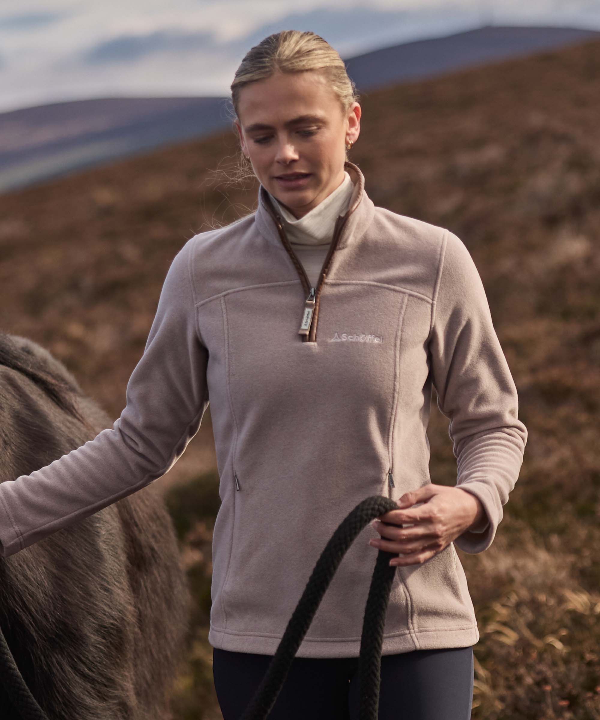 Woman wearing a taupe Schöffel fleece quarter-zip, holding a horse's lead rope on moorland.
