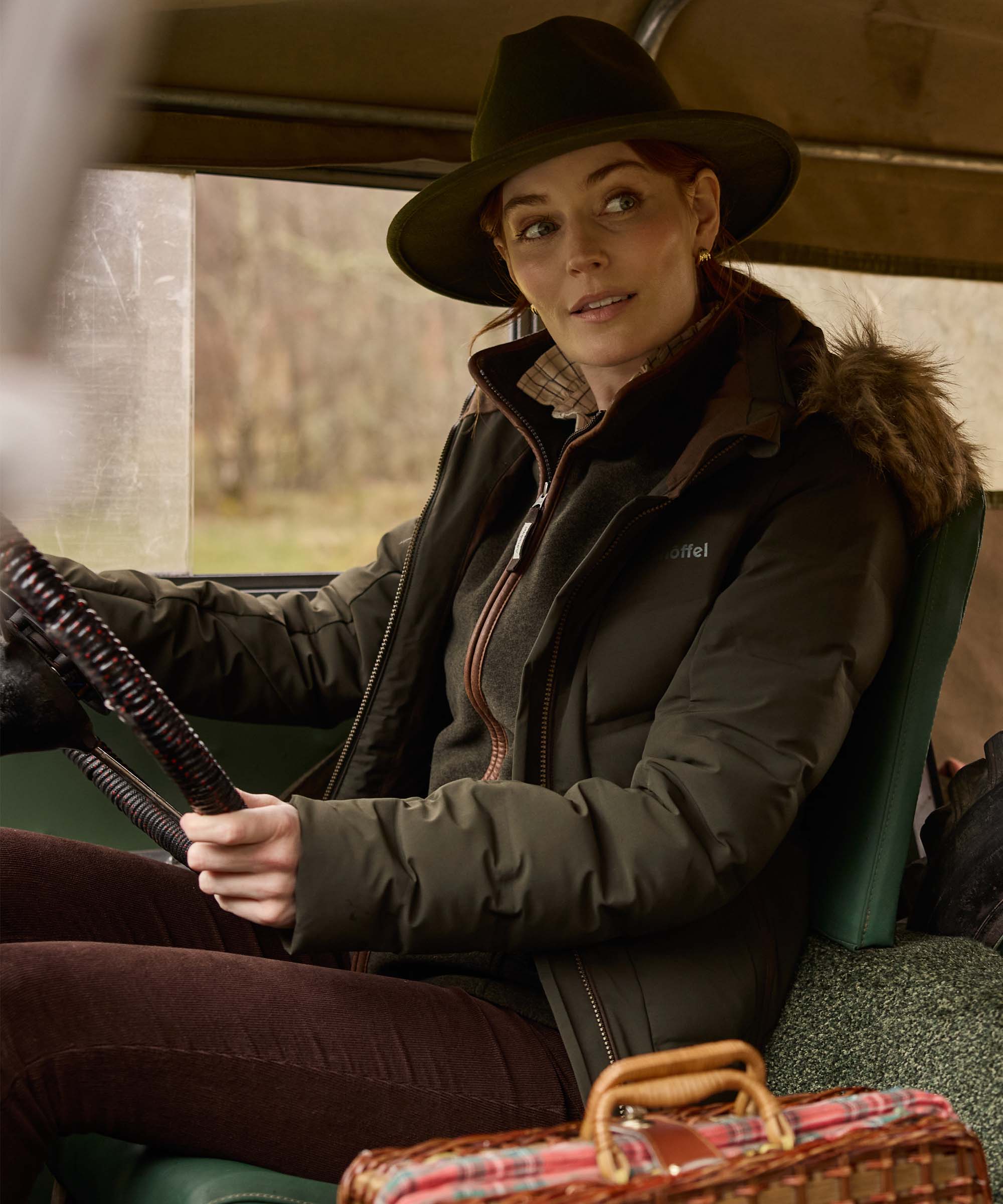 A woman wearing a dark wide-brim hat, a dark brown puffer jacket with faux-fur trim, and brown trousers sits inside a vehicle, holding a steering wheel.