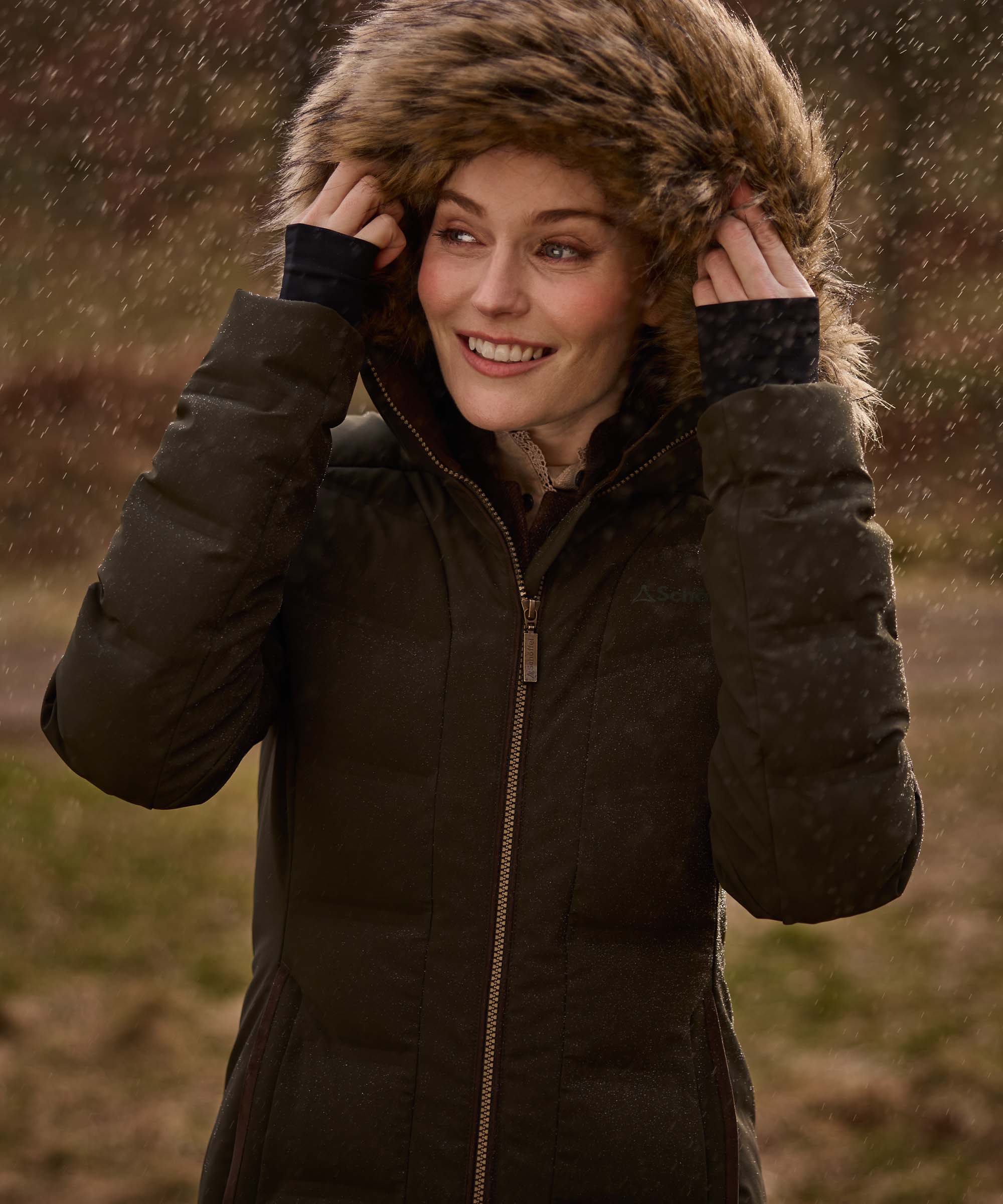 A woman wearing a dark brown puffer parka with a large brown faux-fur trimmed hood, standing outdoors in falling rain.