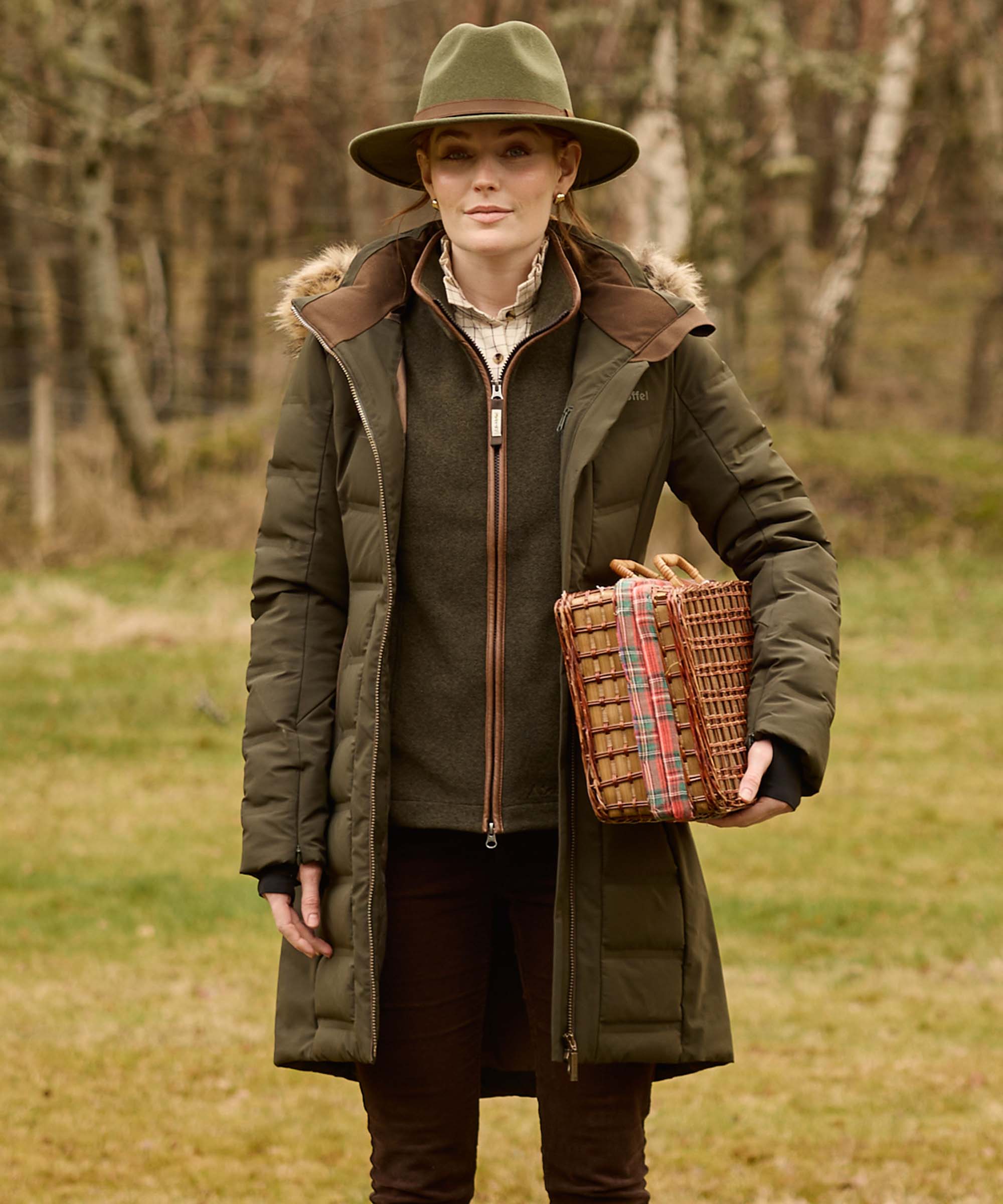 A woman stands outdoors in a wooded setting wearing a long olive green quilted parka with a brown fur-trimmed hood, a dark green jumper, brown trousers, an olive green hat, and holding a woven basket.