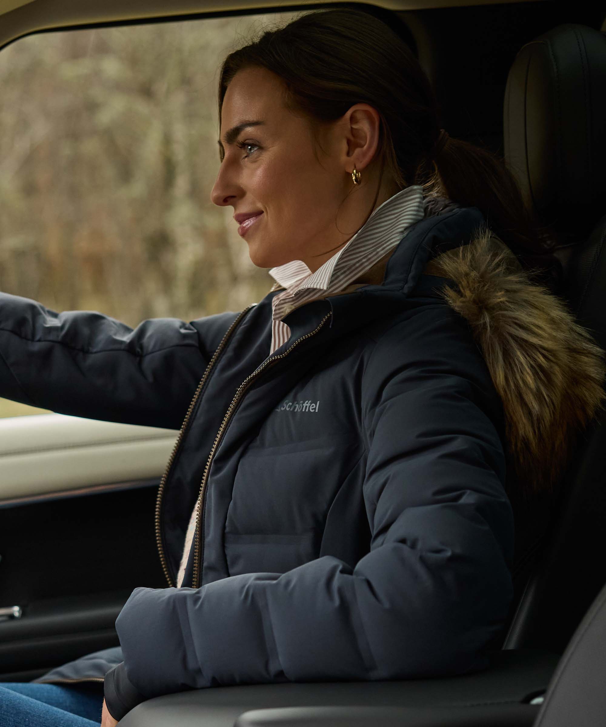 A woman wearing a navy blue puffer jacket with a brown faux-fur trimmed hood sits inside a car, looking out the window.