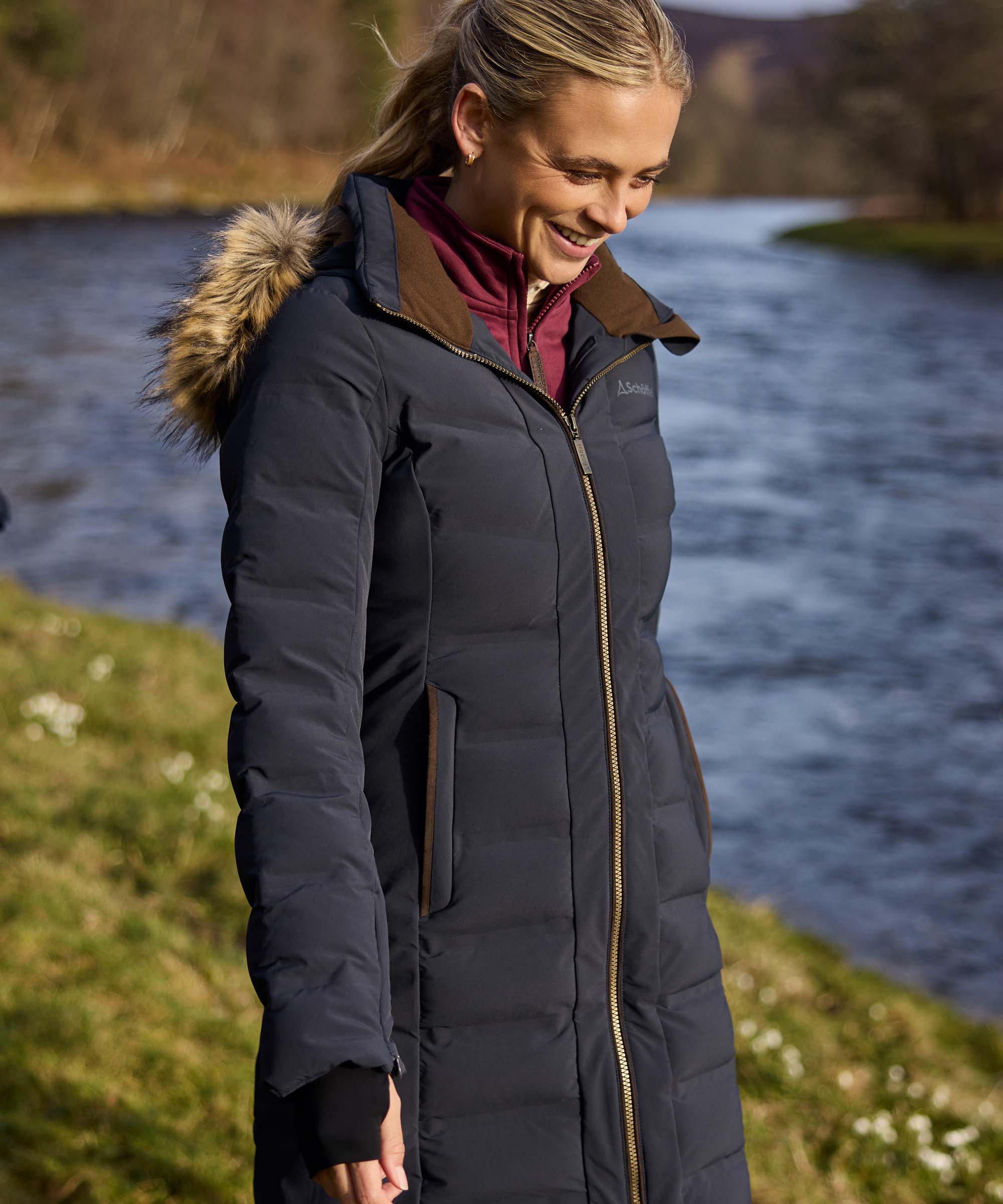 A woman wearing a long dark navy puffer parka with a brown fur-trimmed hood and a gold zipper, standing outdoors near a river.