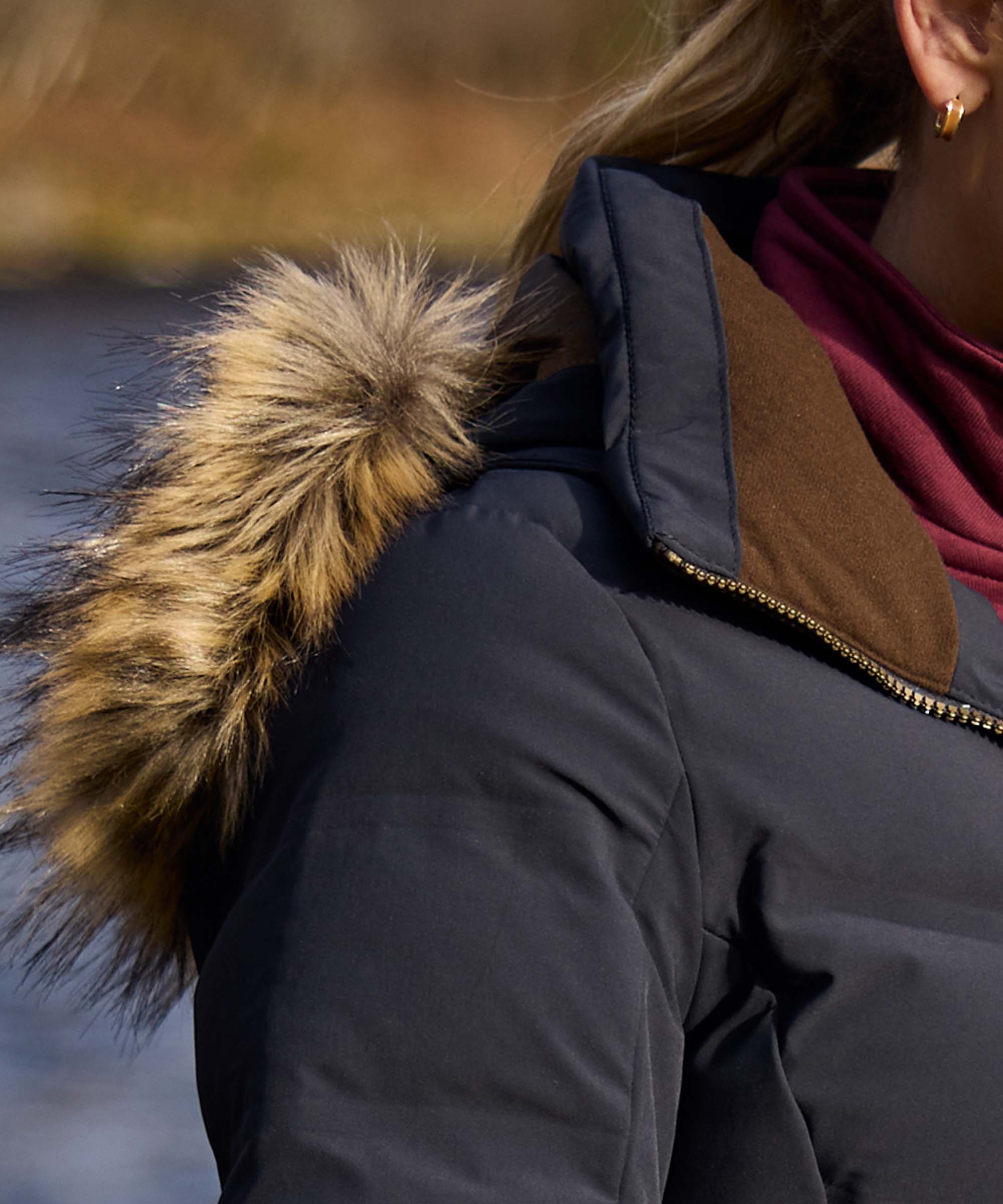 Close-up of a person wearing a dark navy winter jacket with a brown suede-like collar and a fluffy tan and brown faux-fur trim around the hood, outdoors near water.