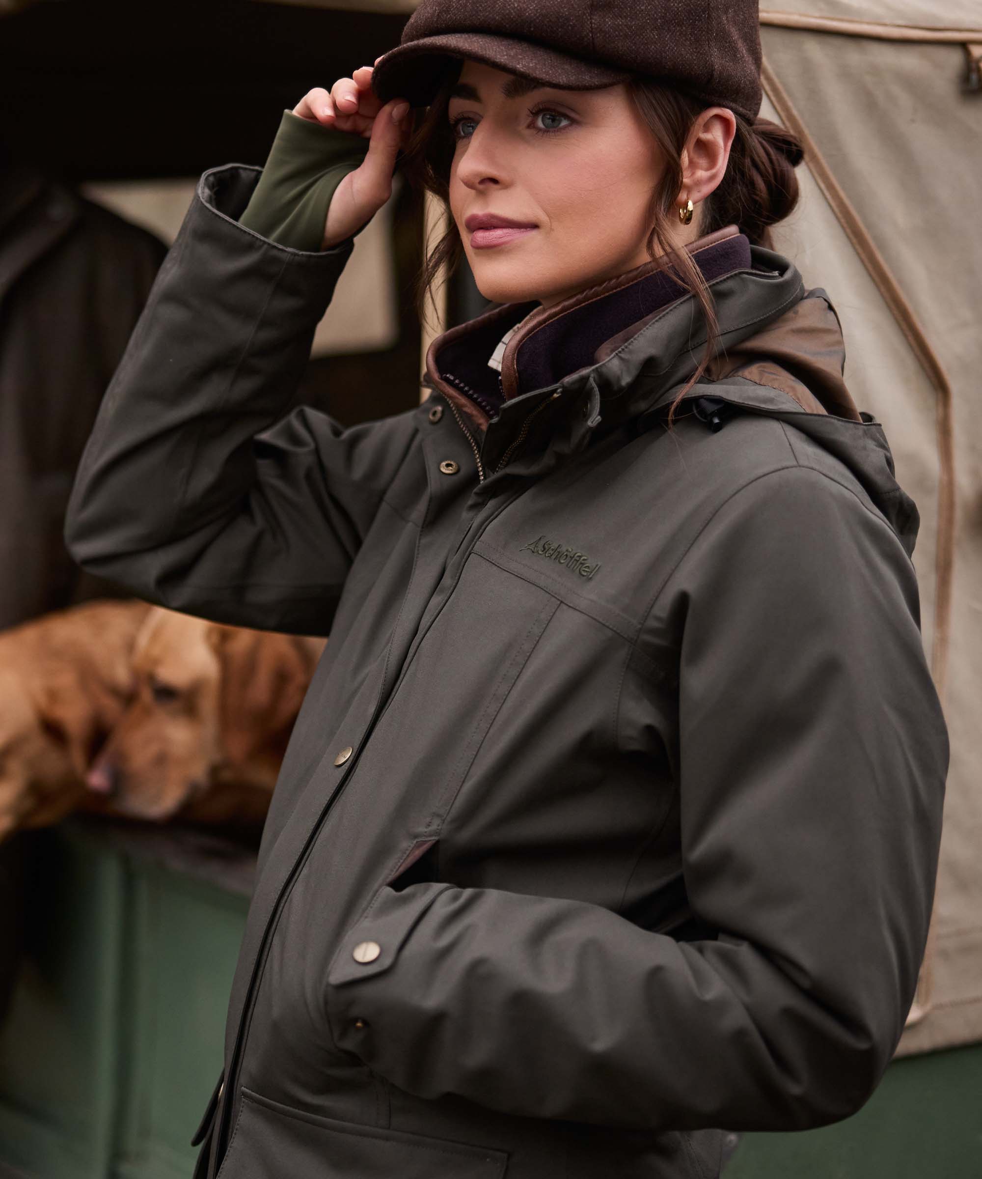 A woman wearing a dark olive green waterproof jacket and a brown flat cap, posing in an outdoor rustic setting with a dog in the background.
