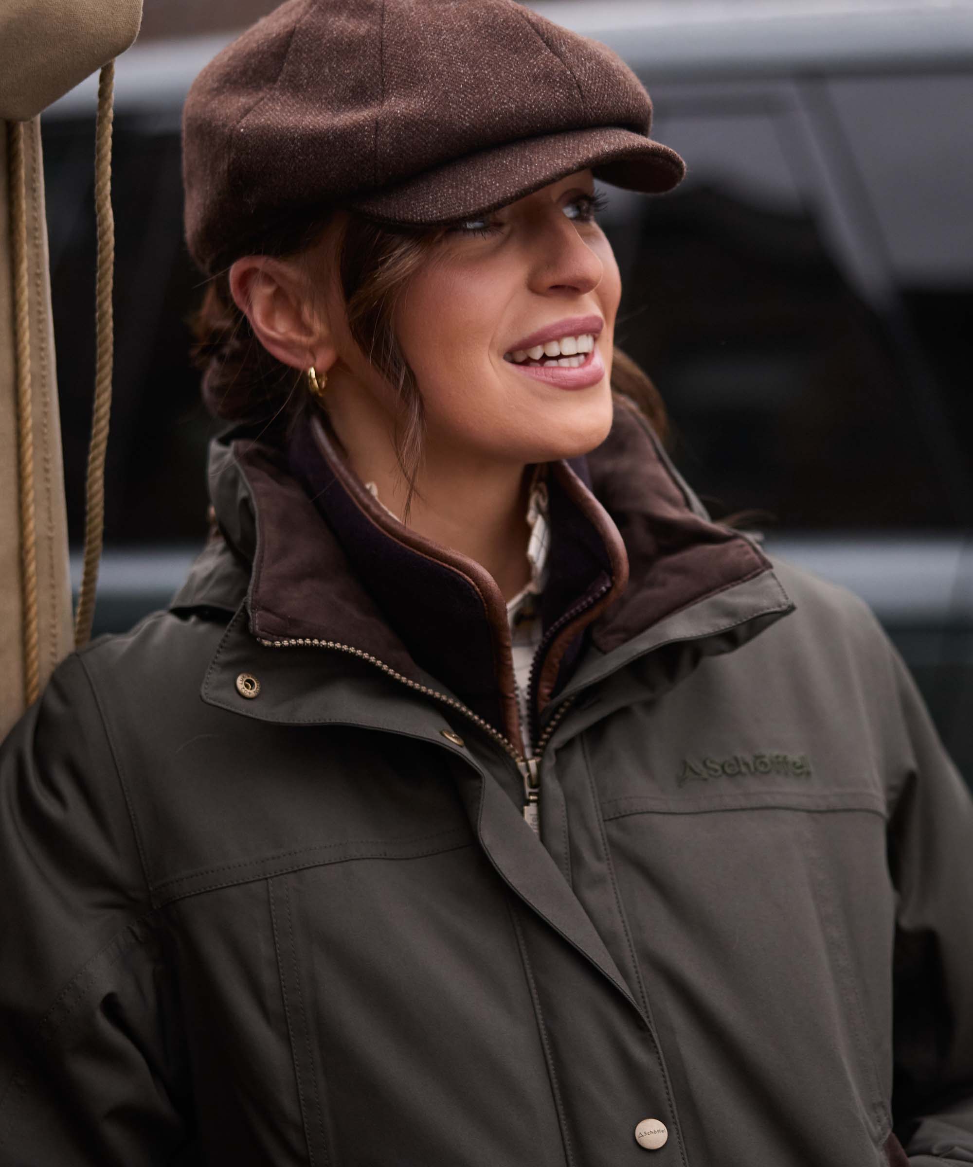 A woman wearing a brown newsboy cap and a dark olive green outdoor jacket with a brown collar, shown in a close-up shot.