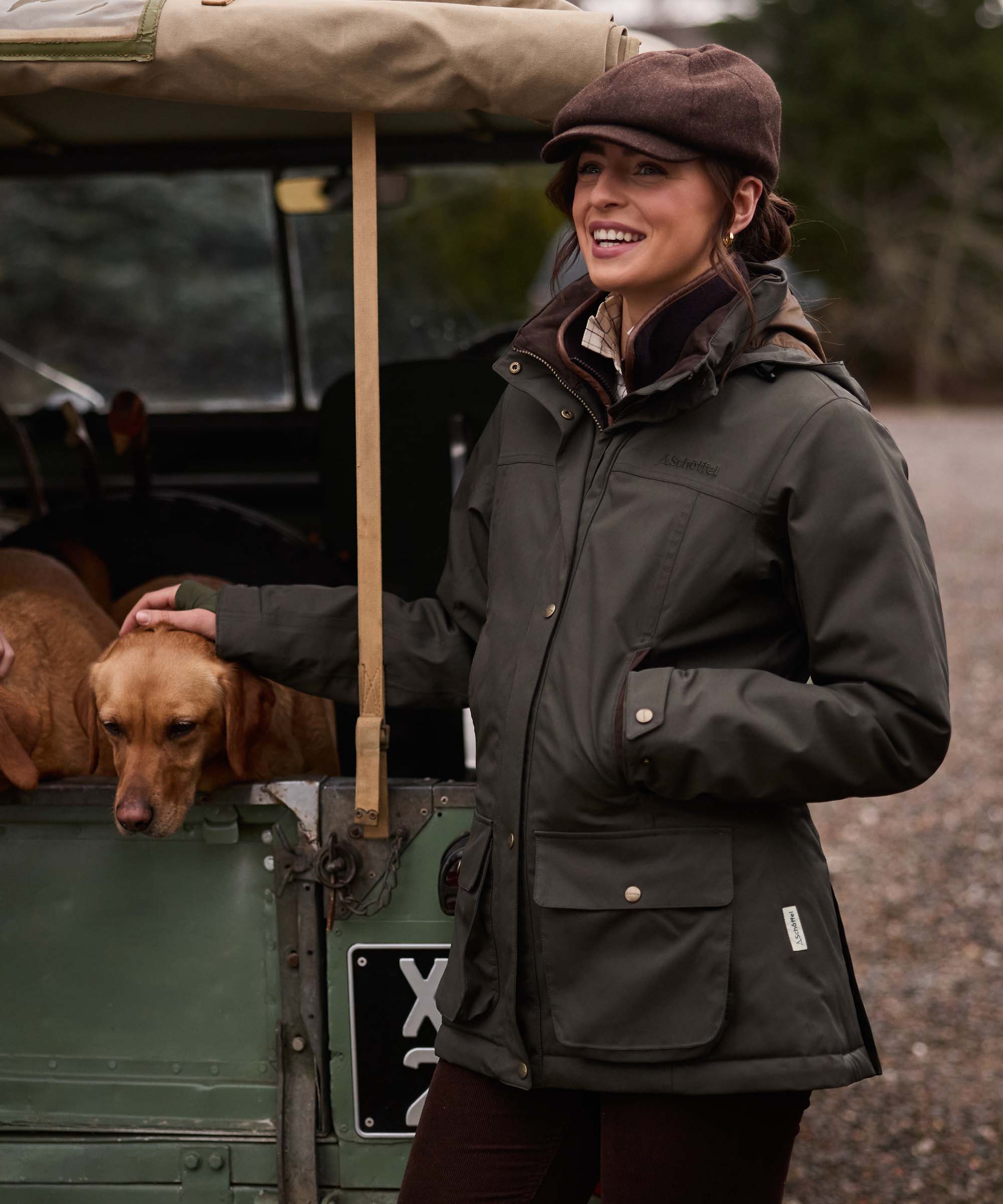 A woman wearing a dark olive green outdoor jacket and brown flat cap stands beside a vintage green vehicle with a brown dog, smiling in an outdoor setting.