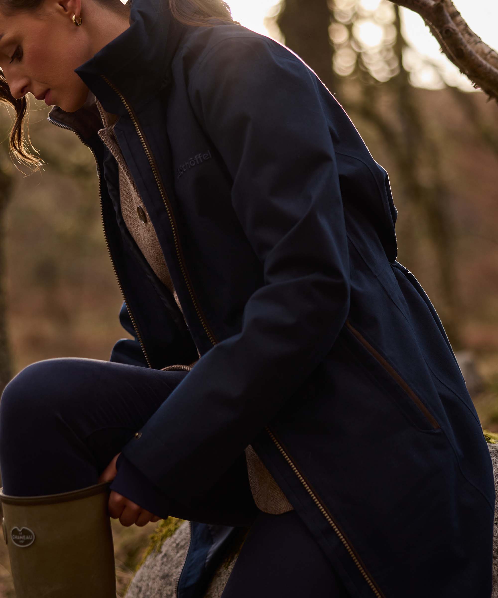 A woman wearing a dark navy hooded jacket, navy pants, and olive green wellies, crouching outdoors in a wooded setting.