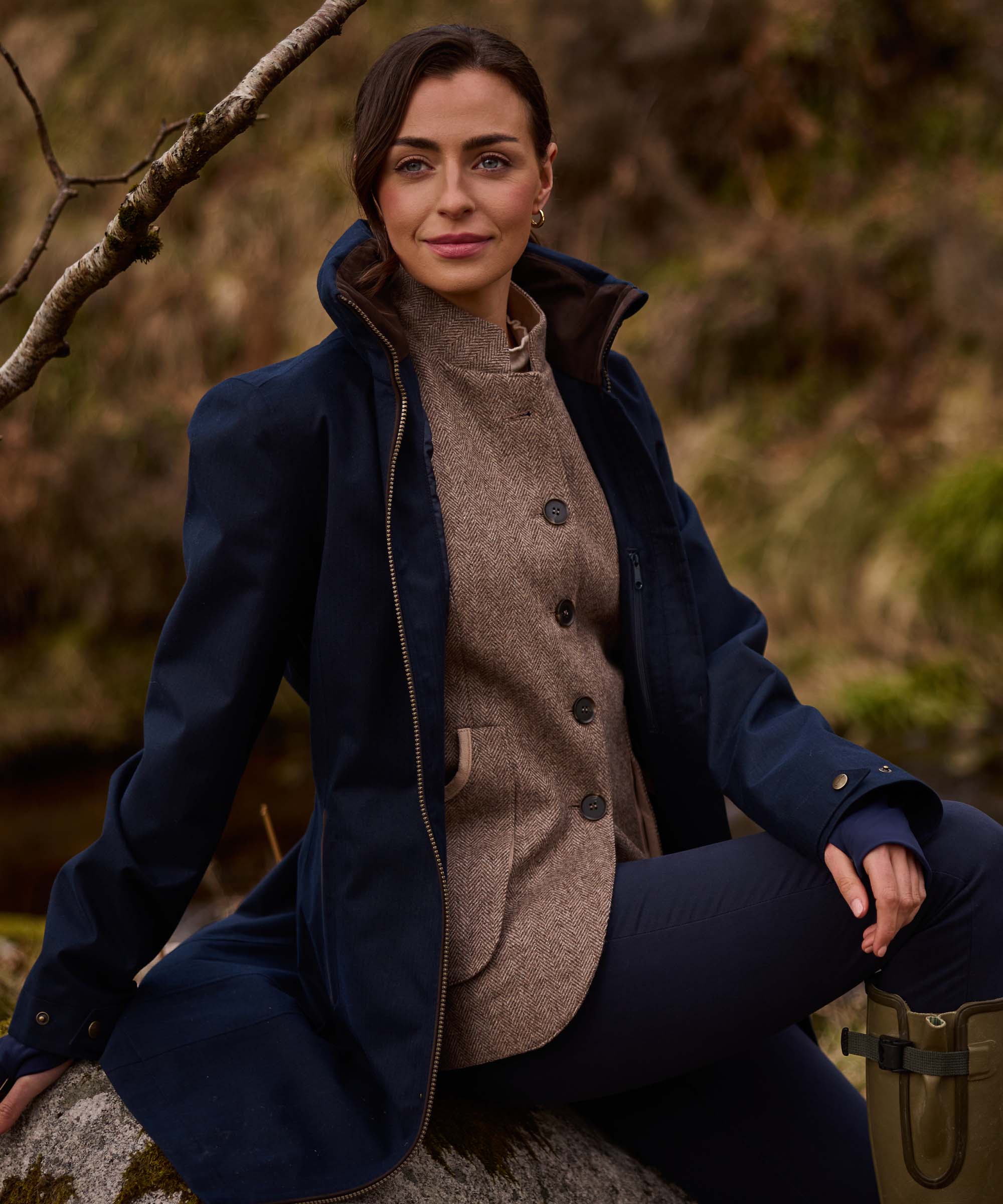A woman wearing a navy blue hooded coat over a brown tweed button-front top and navy trousers, seated outdoors on a rock with a blurred natural background.
