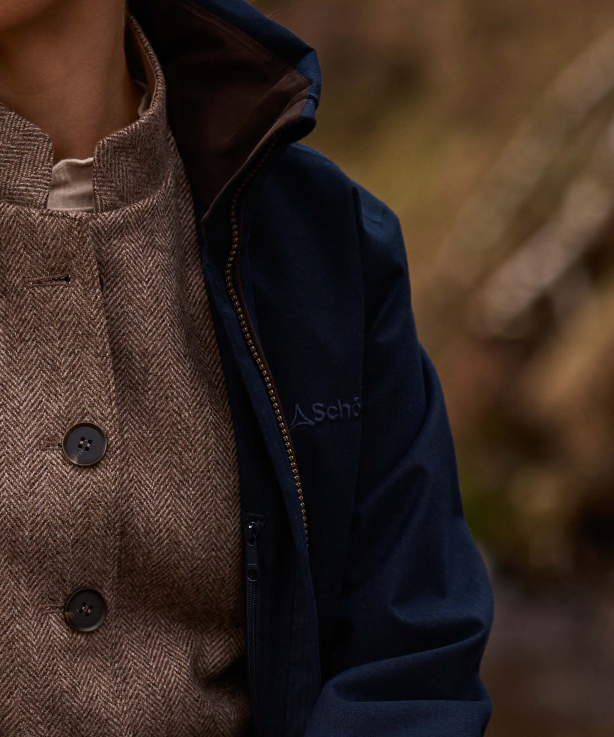 Close-up of a person wearing a navy blue jacket , layered over a brown herringbone tweed button-up waistcoat against a blurred outdoor background.