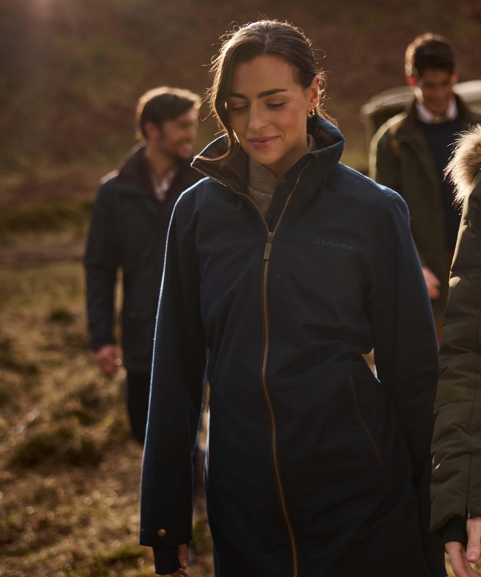 A woman wearing a dark navy hooded jacket with a gold zipper walks outdoors in a grassy field, with other people blurred in the background.