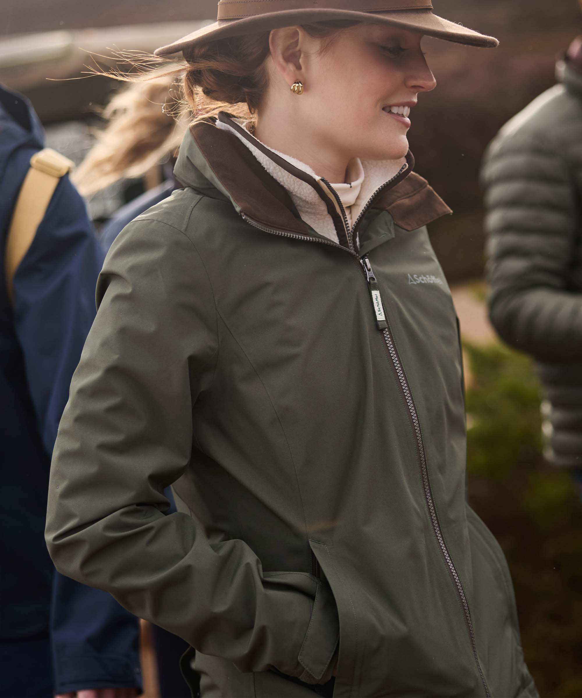 Woman wearing olive green waterproof jacket with brown suede trim and wide-brim hat, smiling outdoors